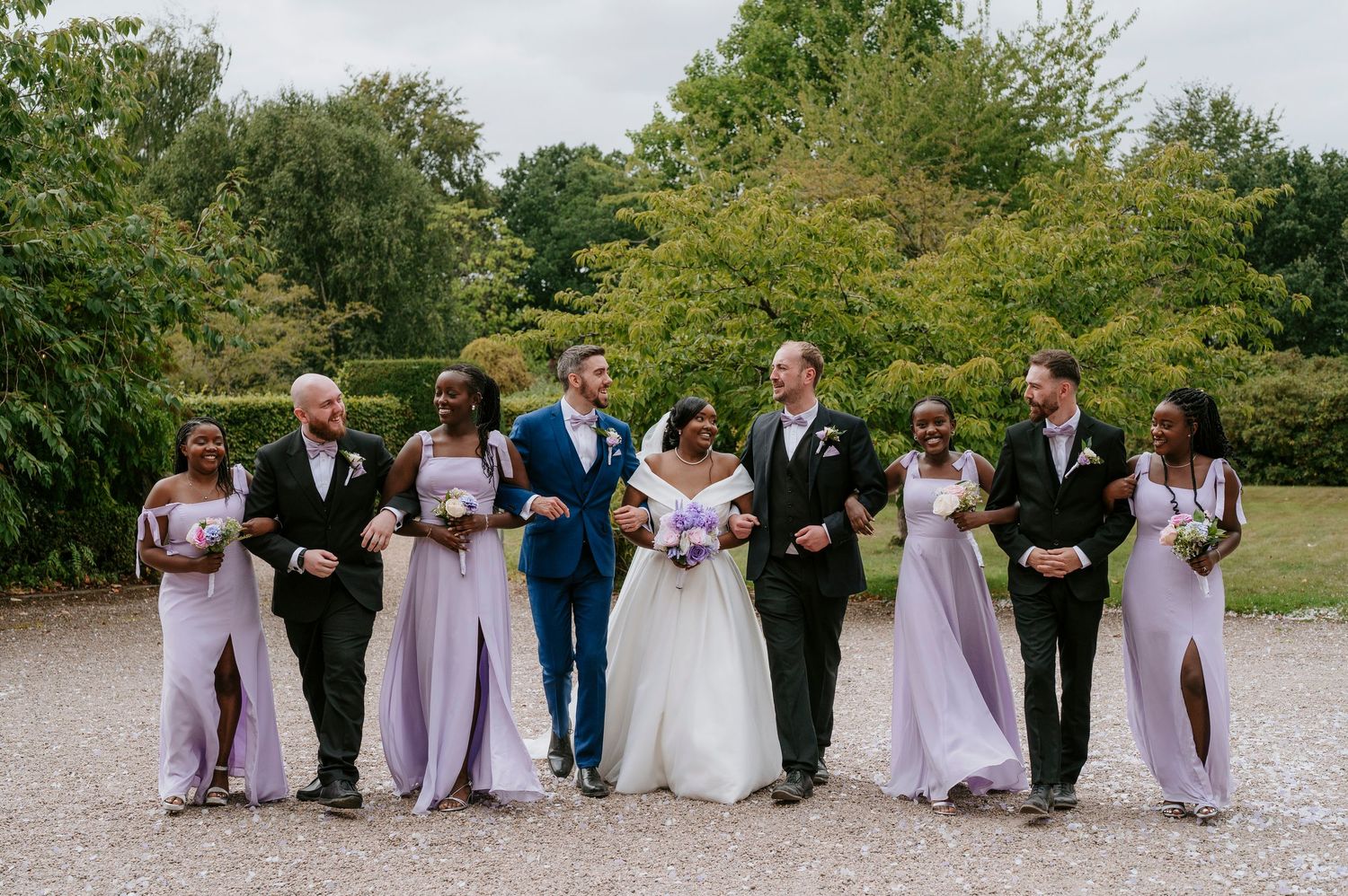 Wedding party poses in a line on a gravel path surrounded by lush green trees and foliage.