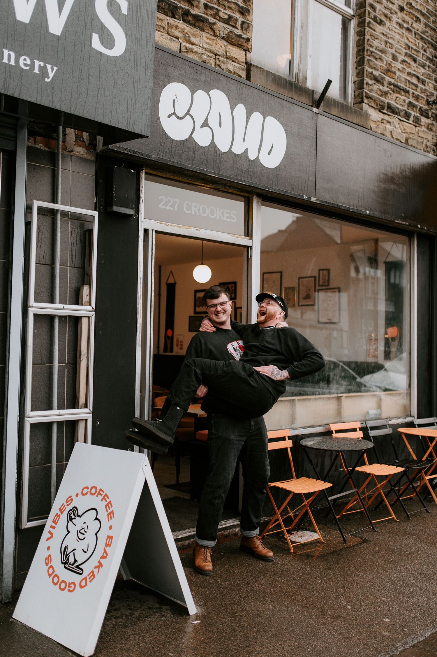 Two people stand outside a cafe storefront with orange chairs and a sandwich board sign on the sidewalk.