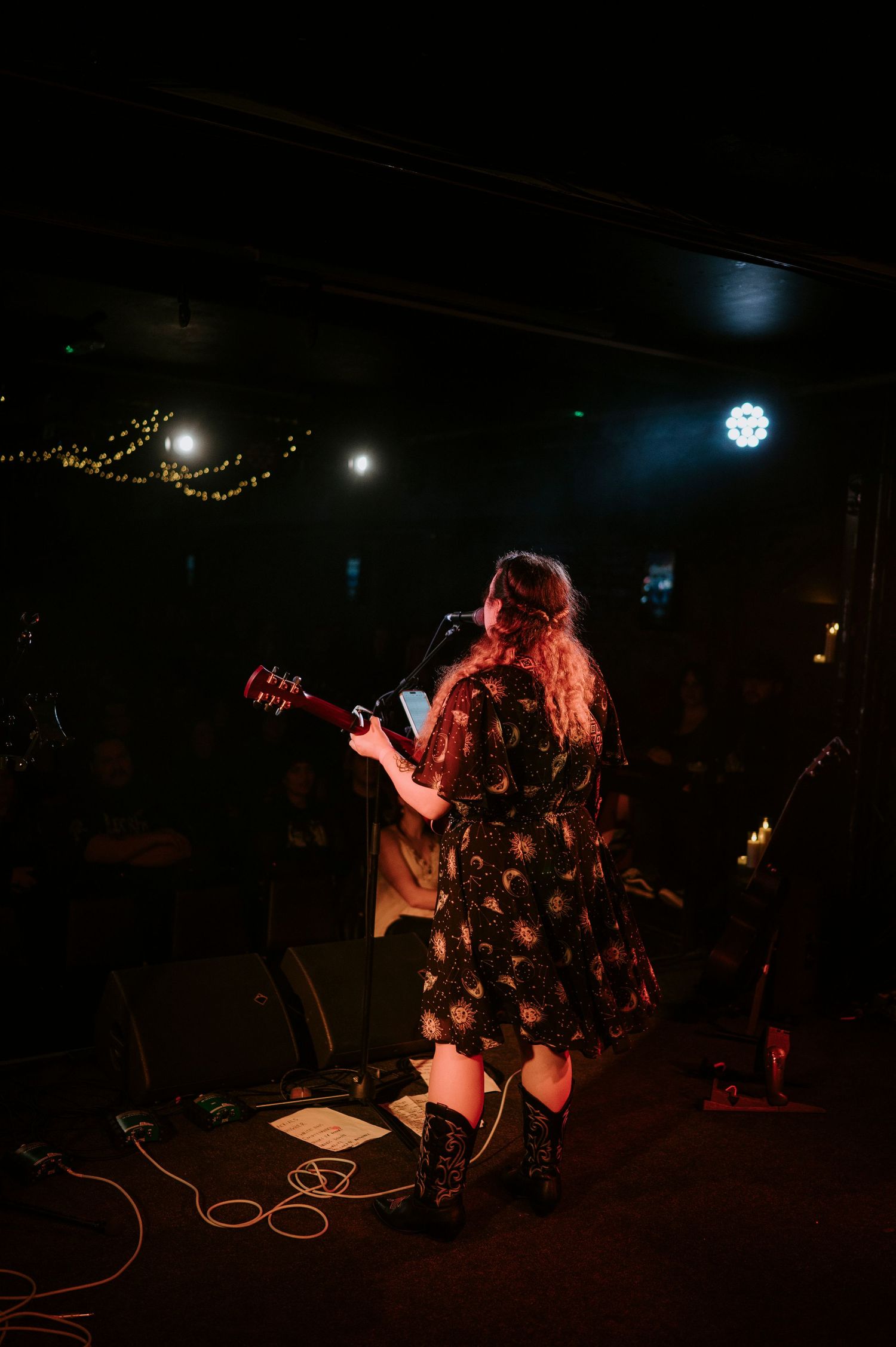 Musician performs on stage in a dark venue with guitar, wearing a floral dress under moody lighting.