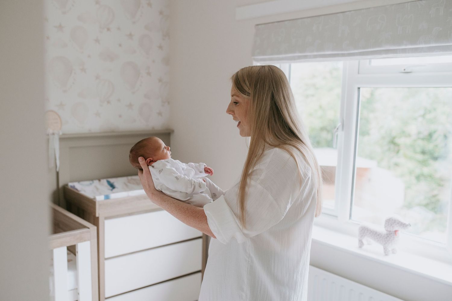 Woman in white dress opens window blinds to let natural light into bright minimalist room.