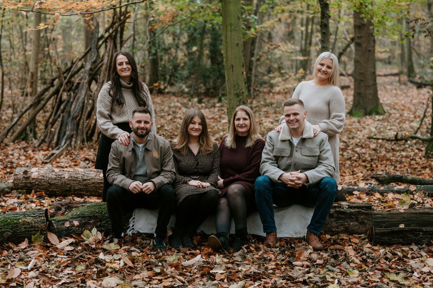 Group sits together on fallen logs in an autumn forest with brown leaves scattered on the ground.