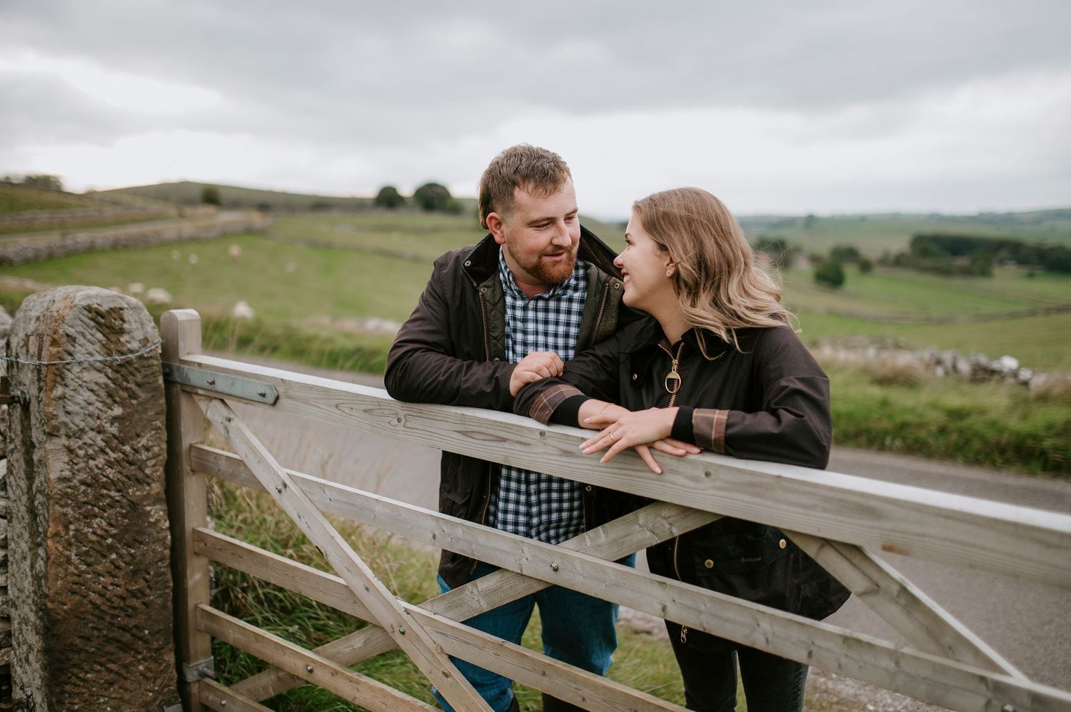 A couple shares a tender moment while leaning on a wooden gate in the rolling countryside on an overcast day.