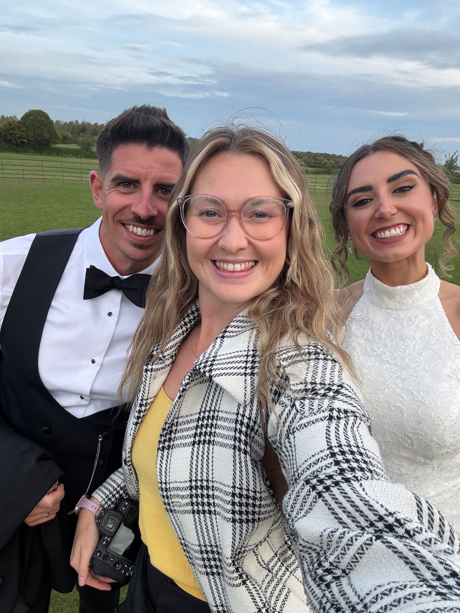 Group of friends in formal attire smiling and taking selfies together in an outdoor field setting.