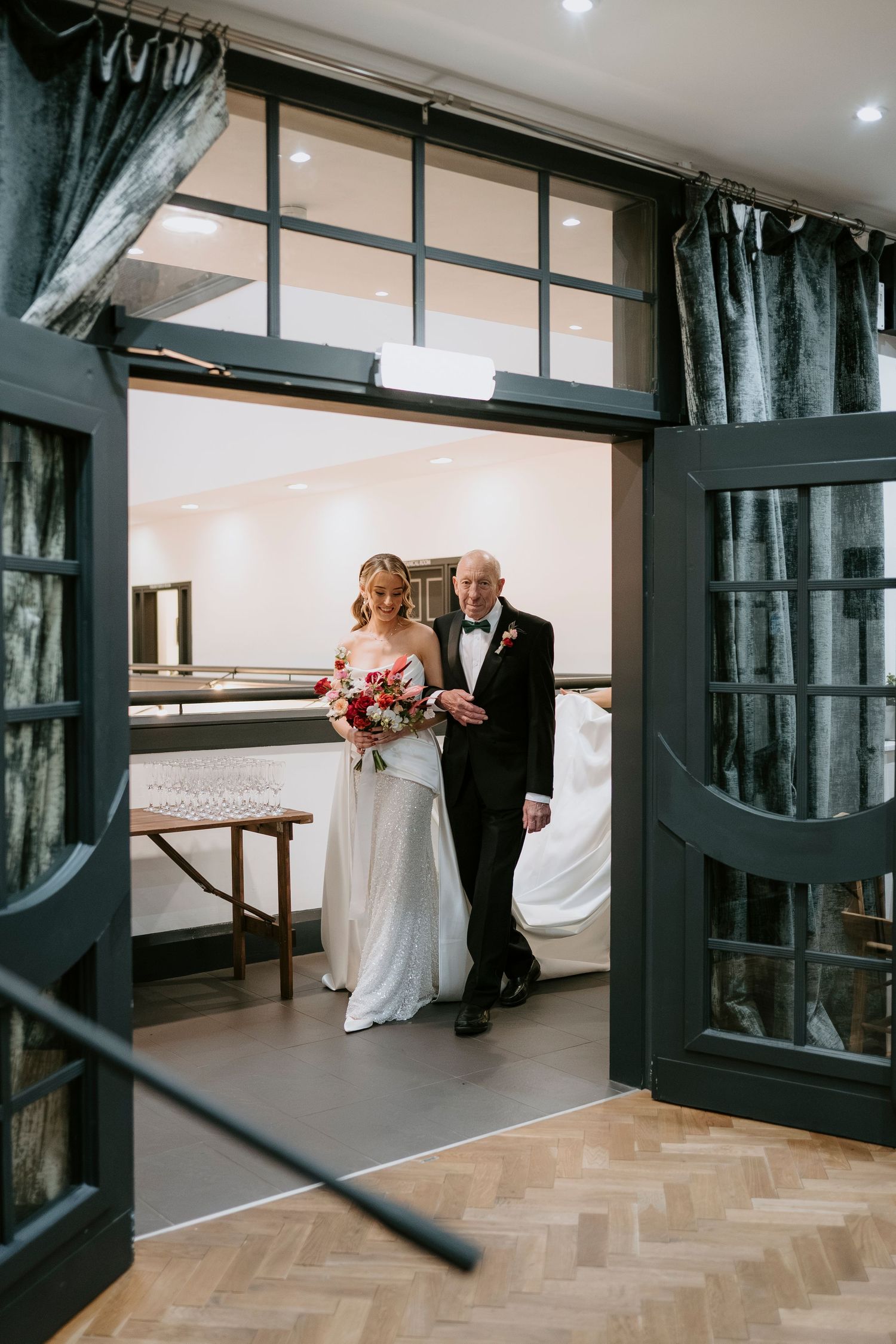 Wedding couple walking through a modern venue with black framed glass doors and herringbone flooring.