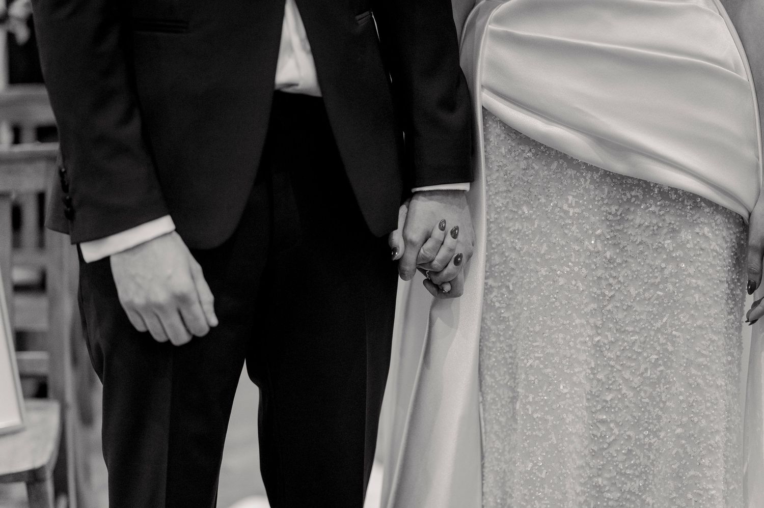 A black and white close-up photograph showing two people holding hands, with formal attire visible.