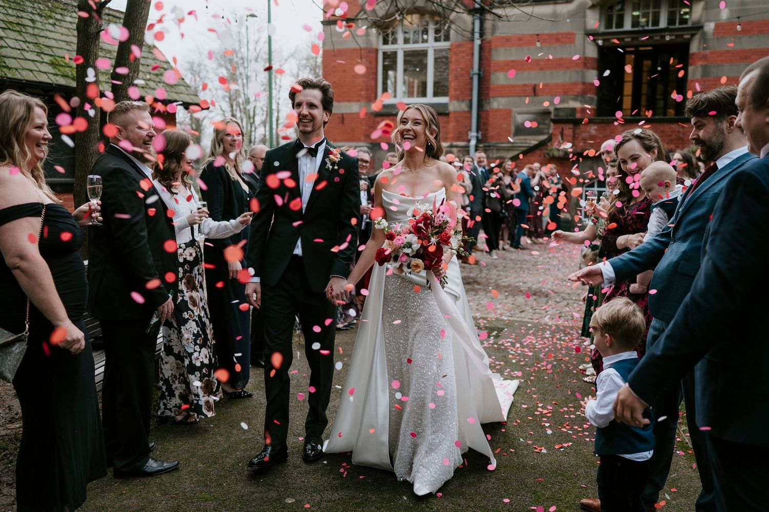 Wedding guests shower newlyweds with pink and white flower petals outside a brick building during celebration.