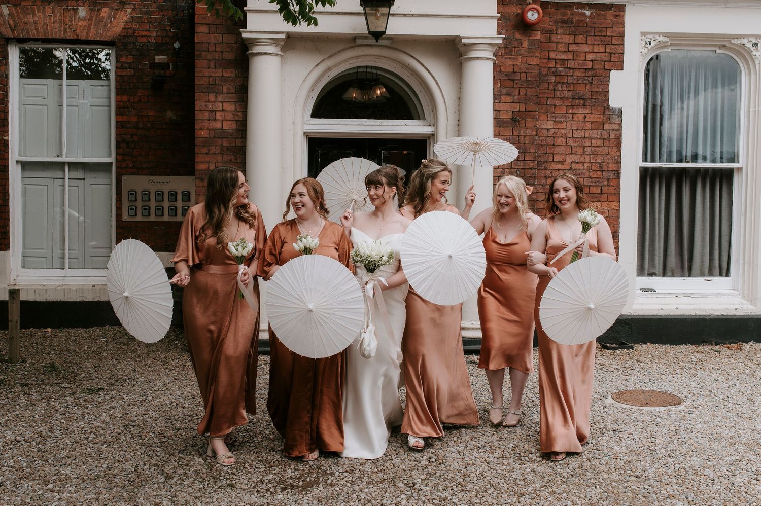 A bridal party in rust-colored dresses poses with white parasols outside a brick building with white columns.