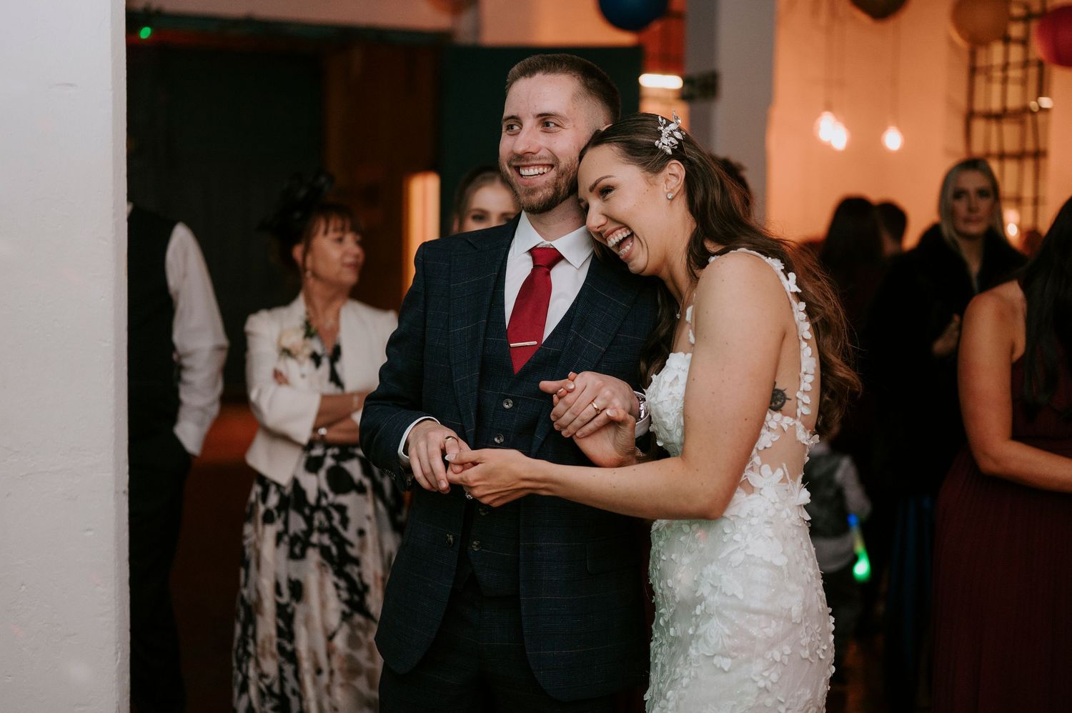 Happy bride and groom laugh together on wedding dance floor surrounded by celebrating guests.