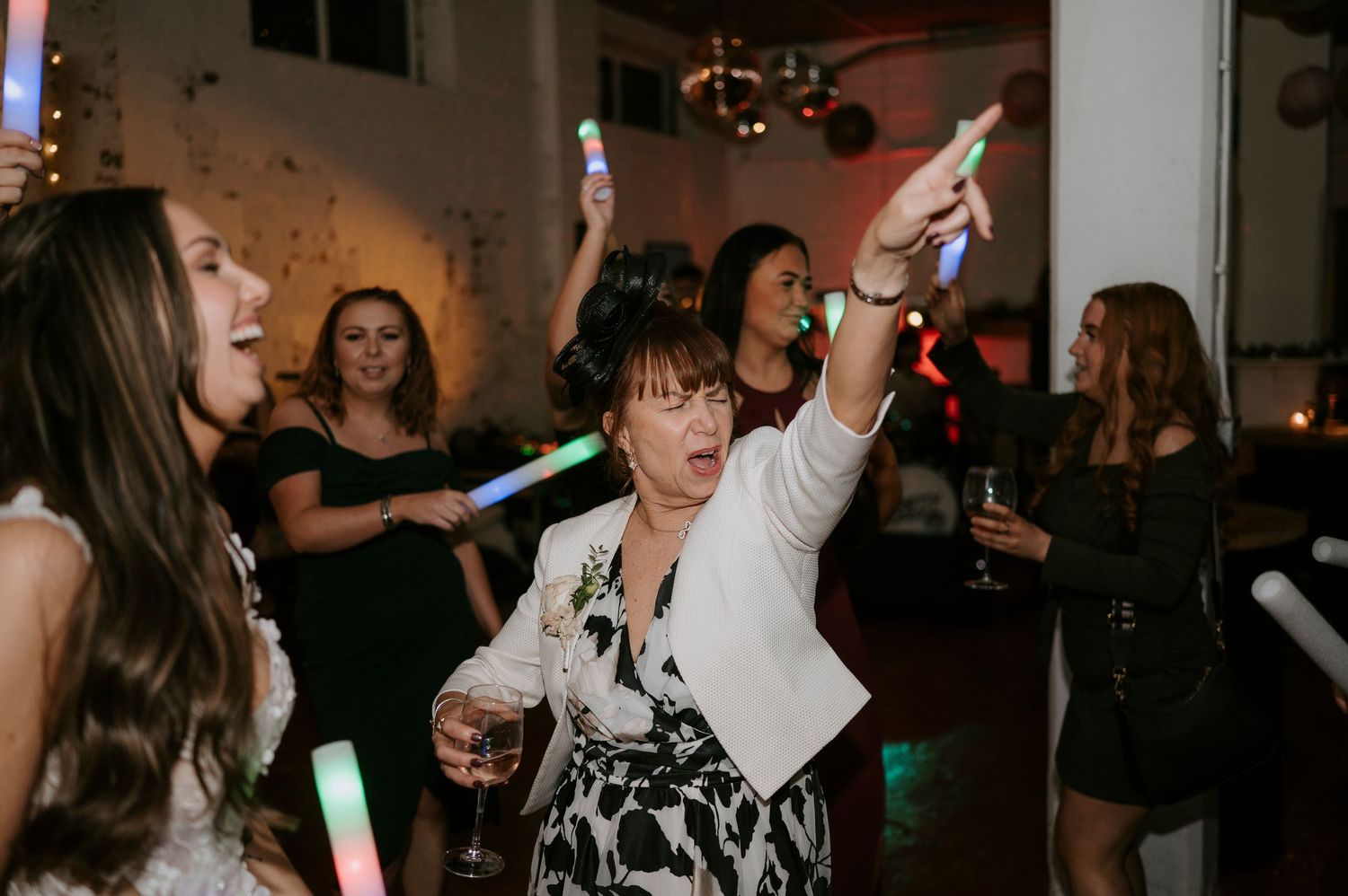 Wedding guests dancing with glow sticks and drinks under red lights at lively wedding reception.
