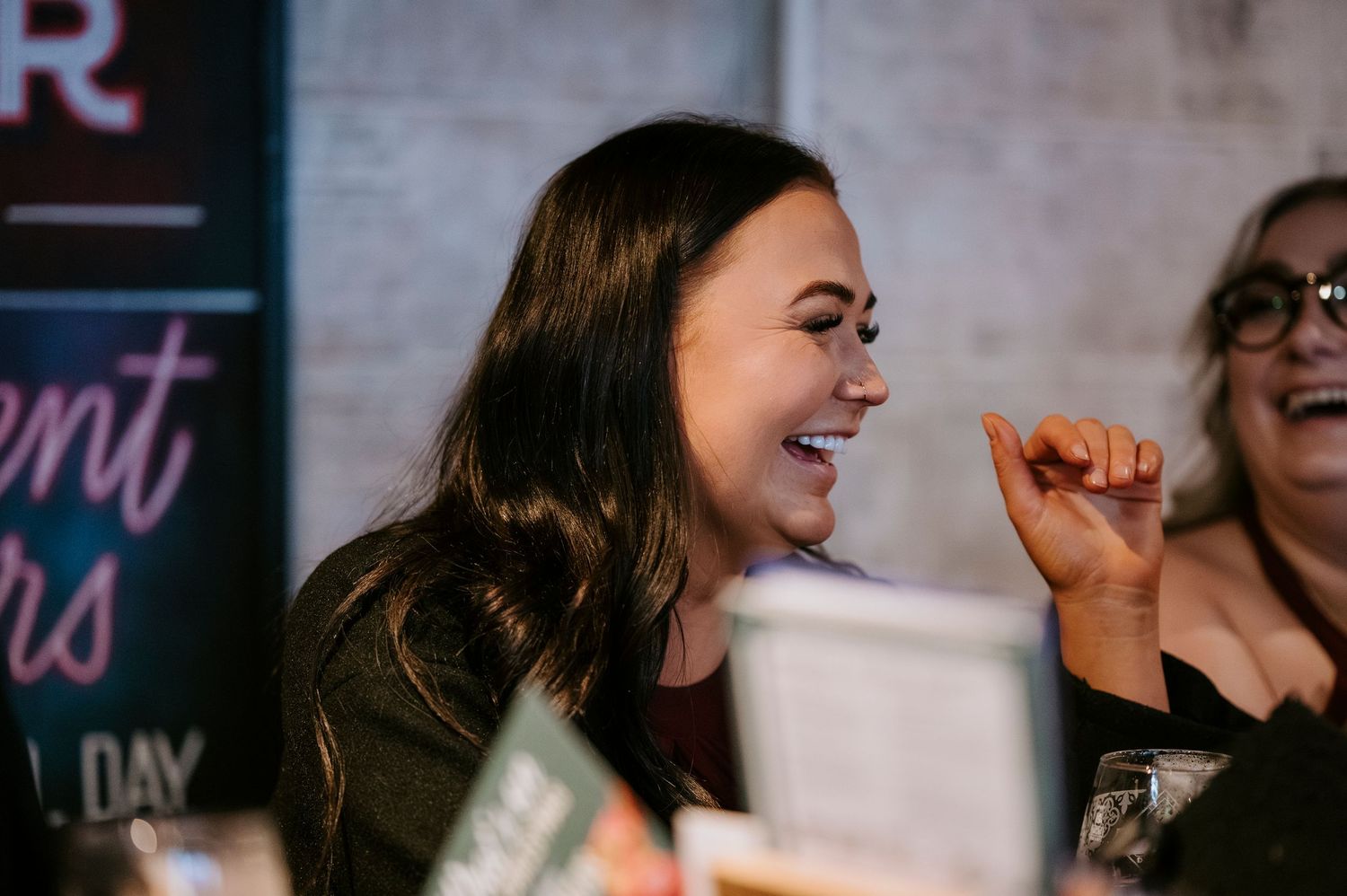 A young woman laughs joyfully at an event, pointing her finger, with another woman visible in the background.