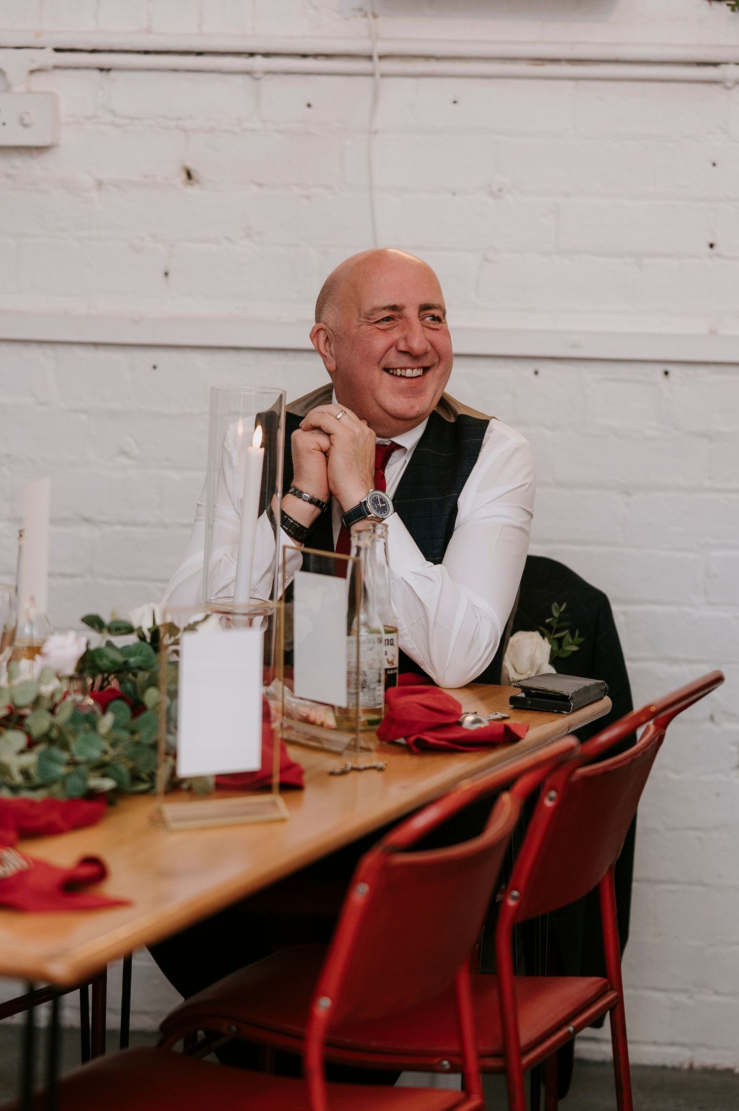 Smiling bald man in white shirt and vest sits at decorated wedding table with red napkins and floral centerpiece.