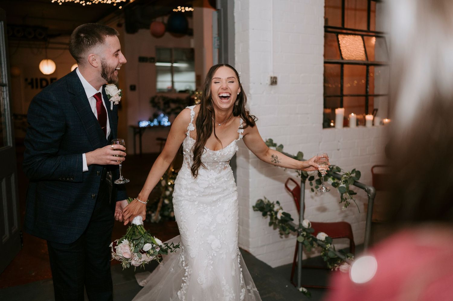 Laughing bride in lace gown and groom in navy suit joyfully enter wedding reception holding bouquet and champagne.