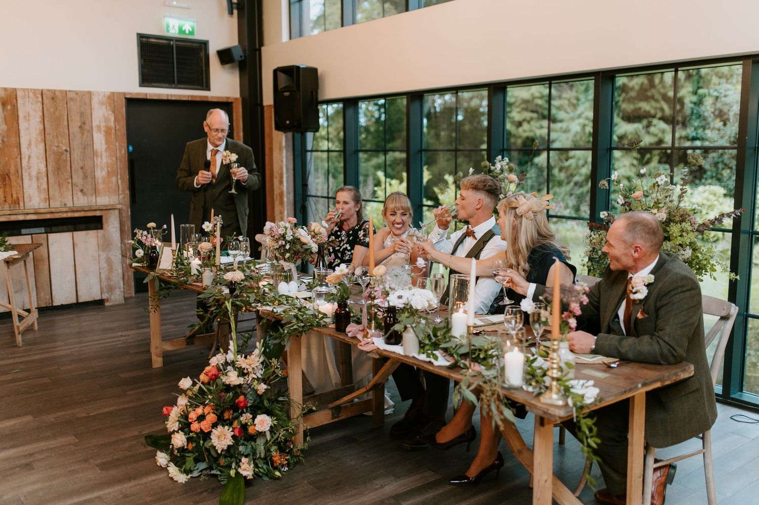 Wedding guests enjoy an elegant reception in a bright, window-filled venue decorated with floral arrangements.