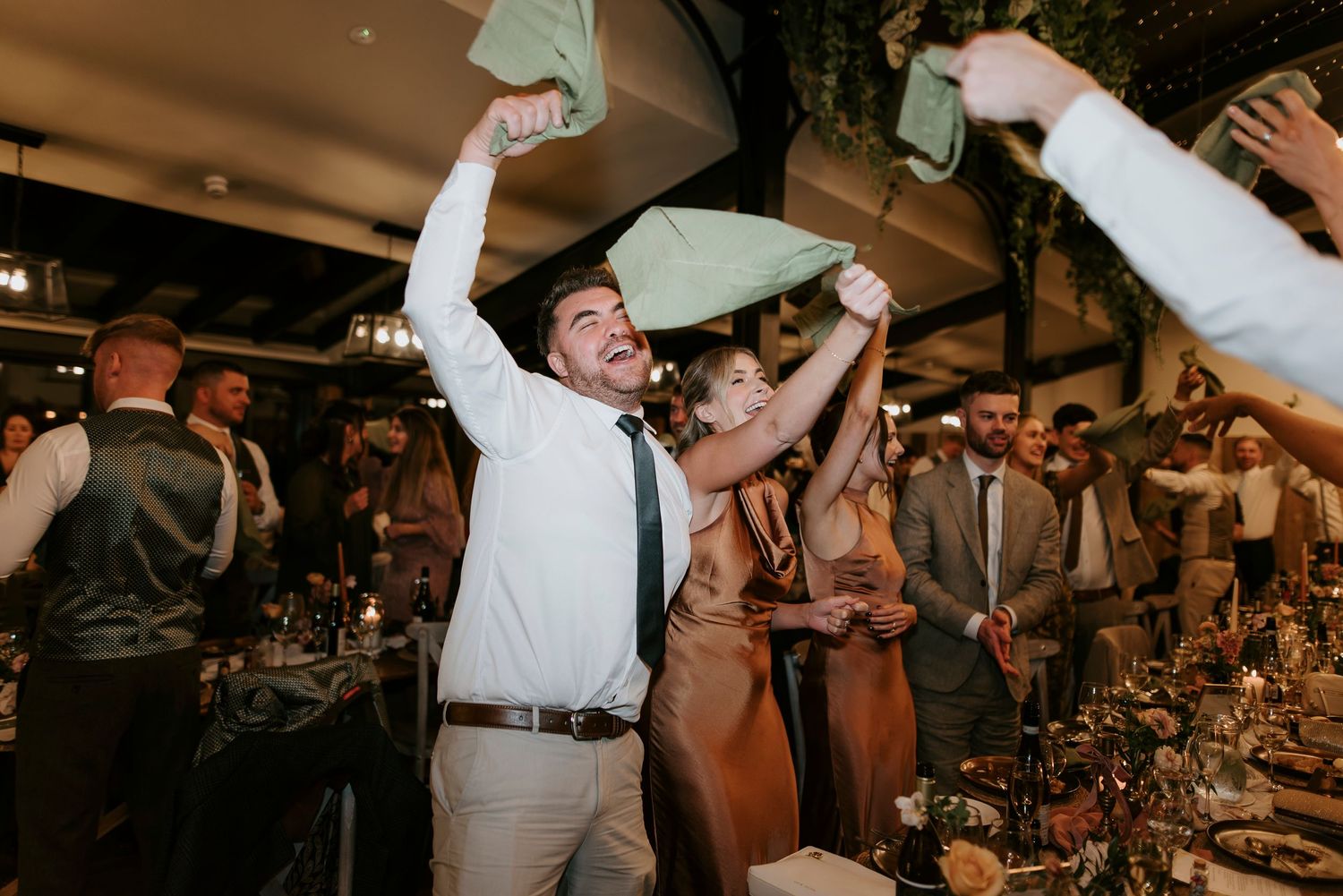 Wedding guests dance and celebrate with raised arms on a dimly lit dance floor during a reception party.