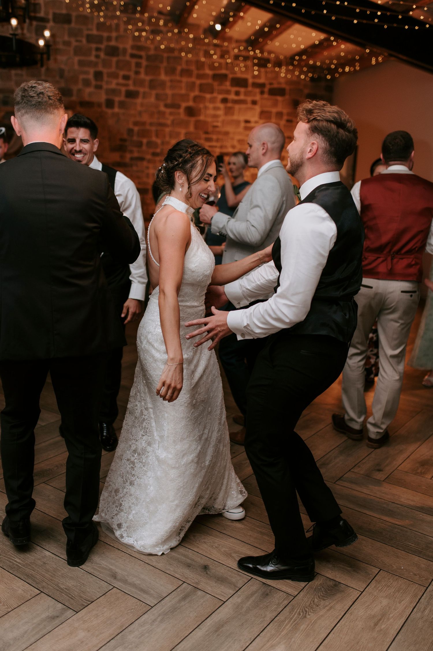 Wedding guests dancing together on a wooden dance floor in a rustic brick-walled venue during the reception celebration.