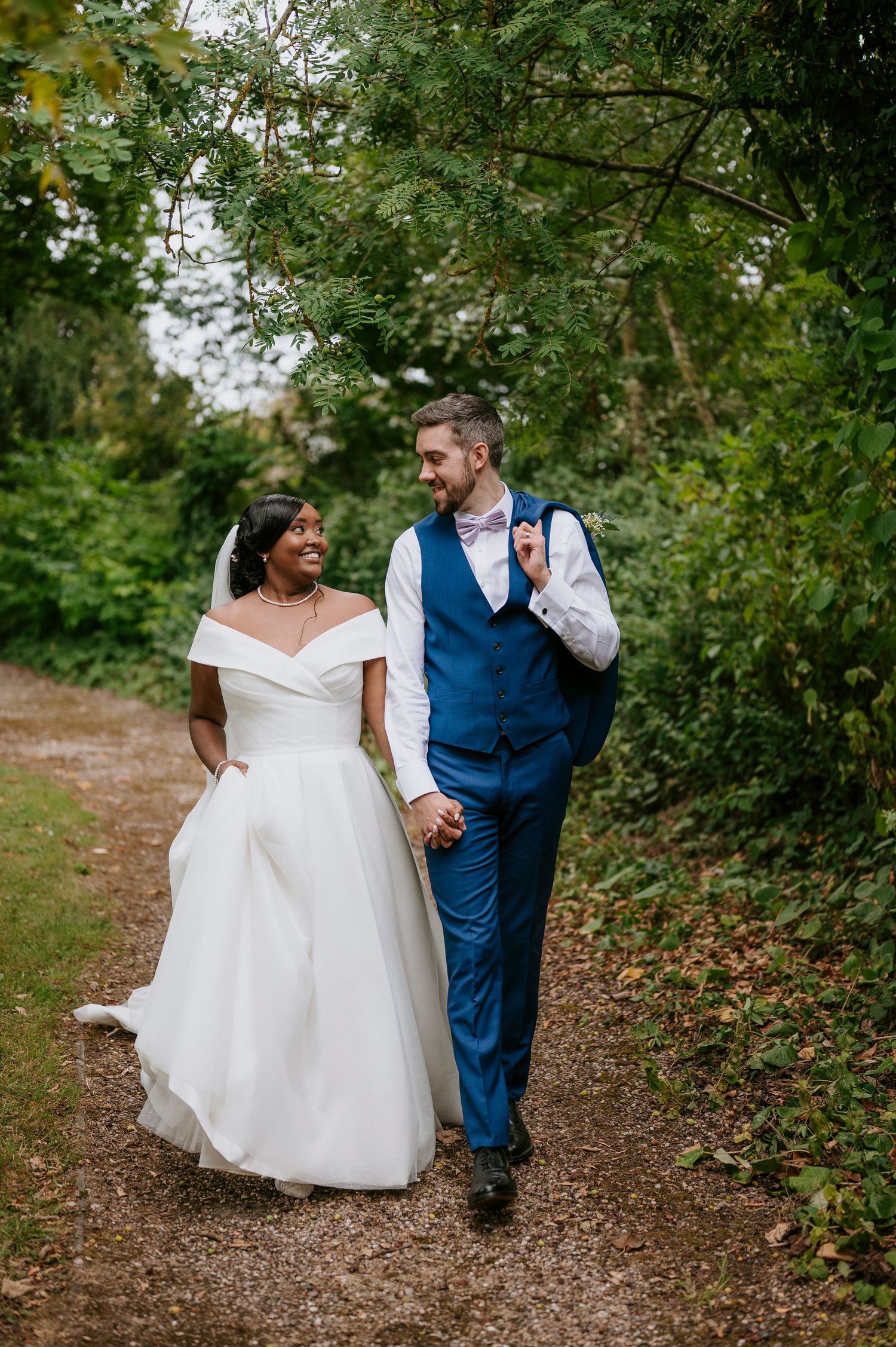 A couple in wedding attire walks along a wooded path together on their special day.