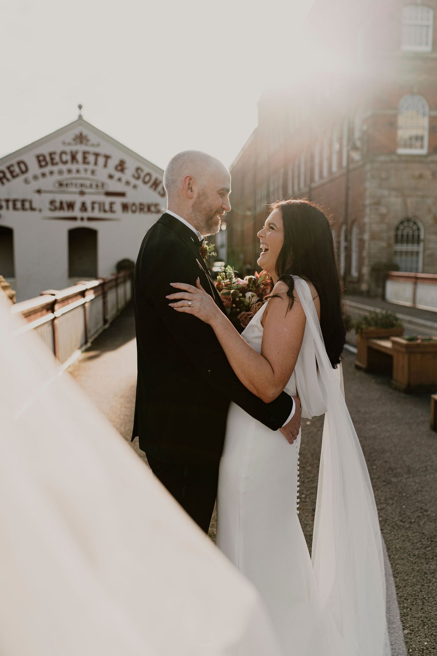 Romantic wedding couple embraces against industrial brick backdrop at sunset.