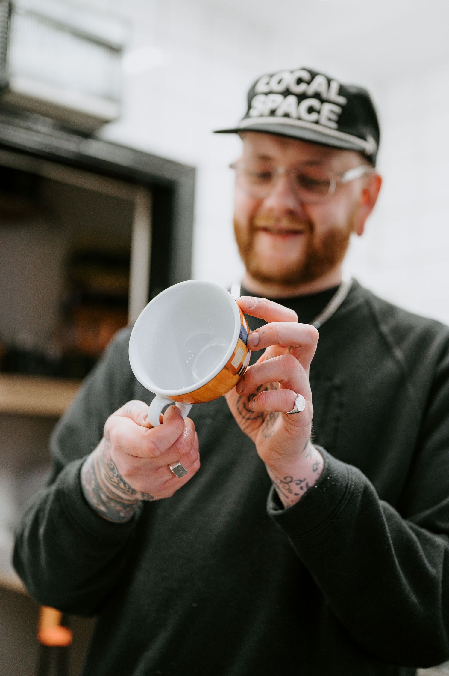 A person in a black sweater holds a teacup while smiling, showing multiple hand tattoos.