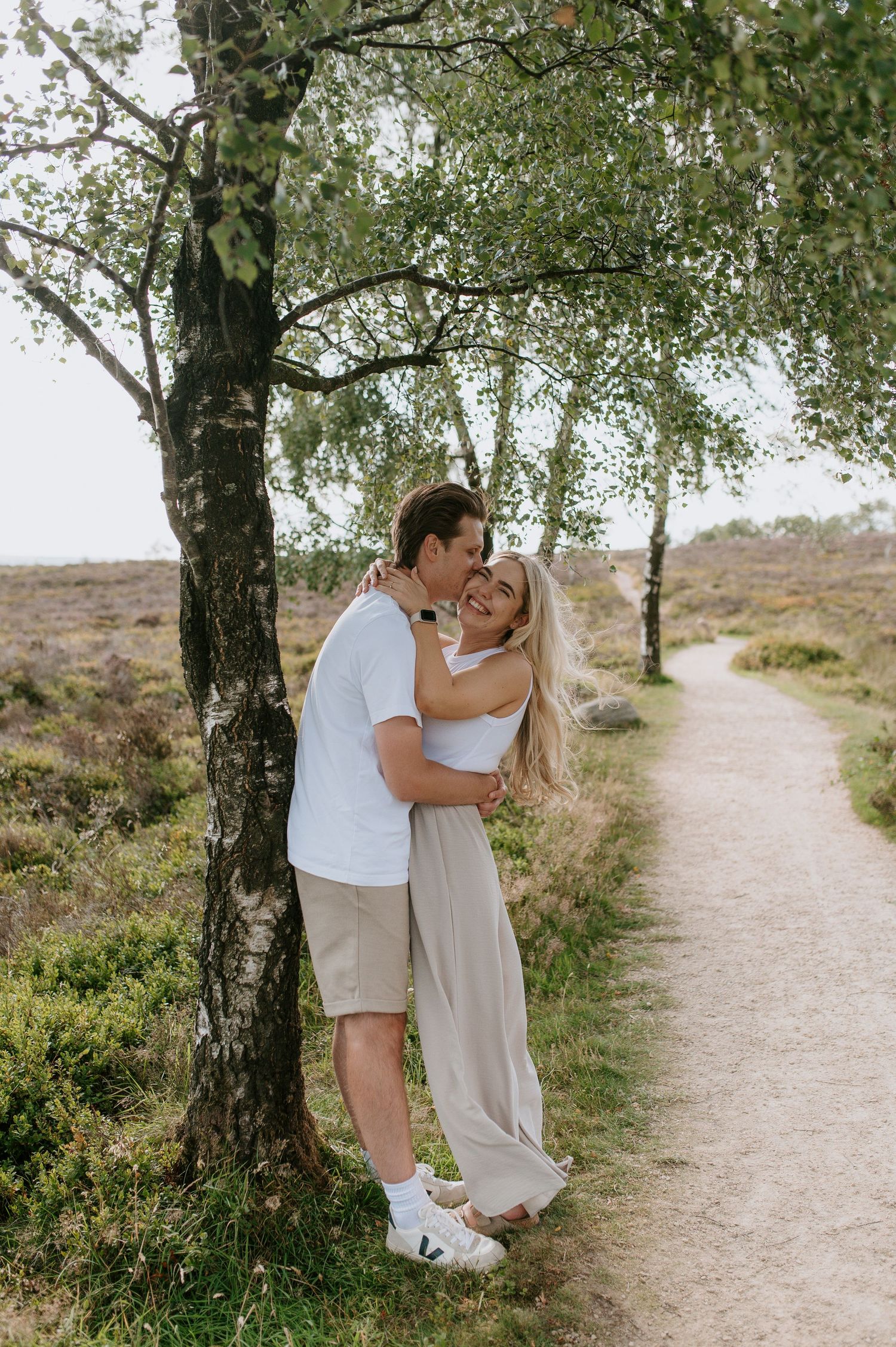 A couple shares an intimate moment on a scenic dirt path surrounded by trees and natural landscape.