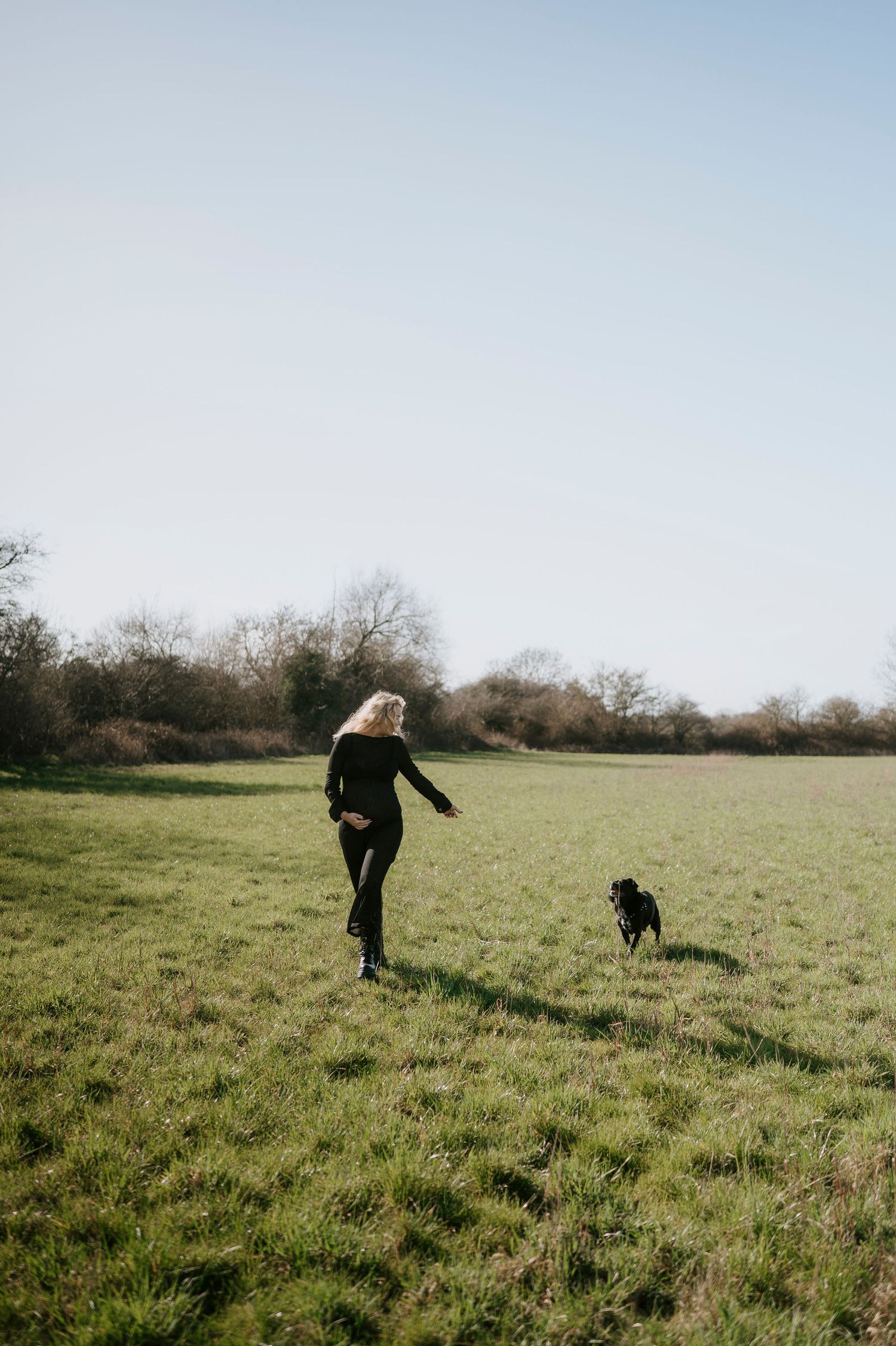 A person in dark clothing runs through a grassy field with a black dog.
