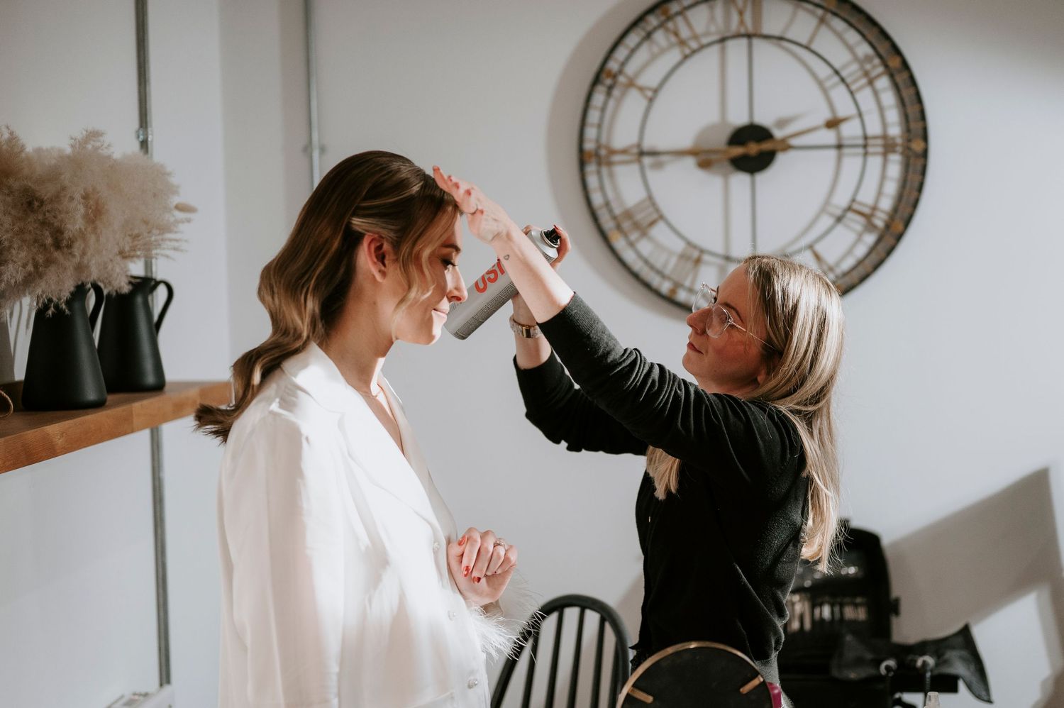 A stylist works on a bride's hair during wedding preparations with a large clock and dried flowers in the background.