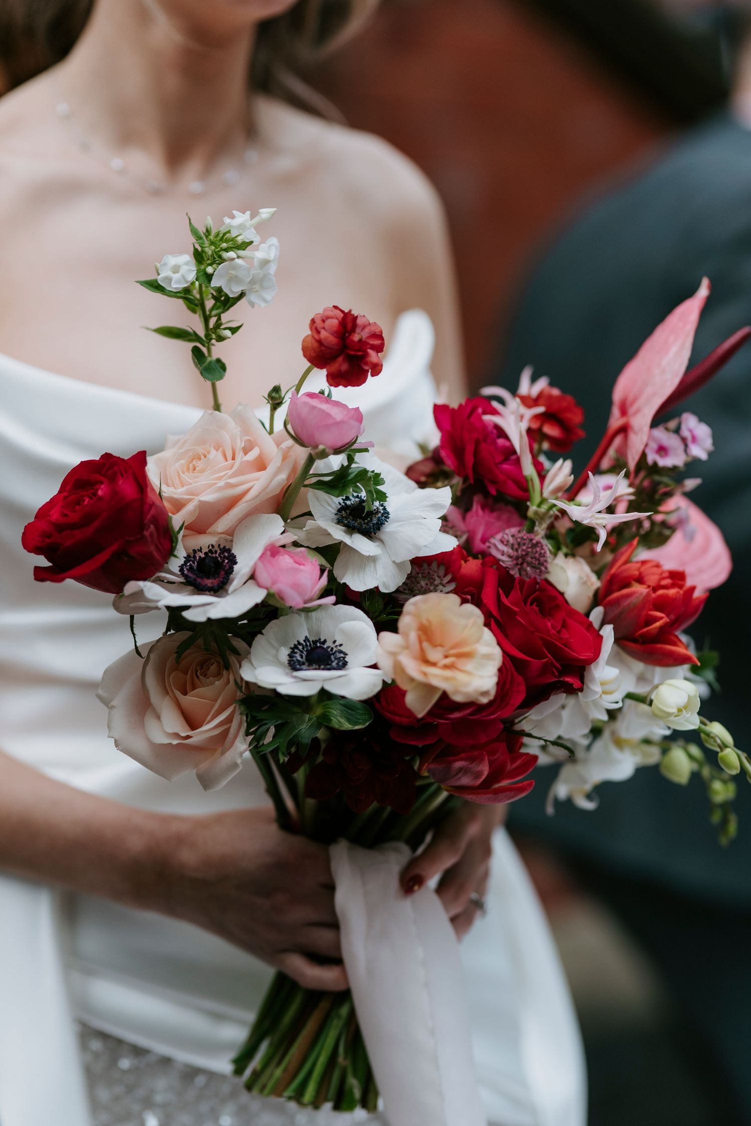 Romantic wedding bouquet features red roses, pink anemones, and blush flowers in a lush garden-style arrangement.