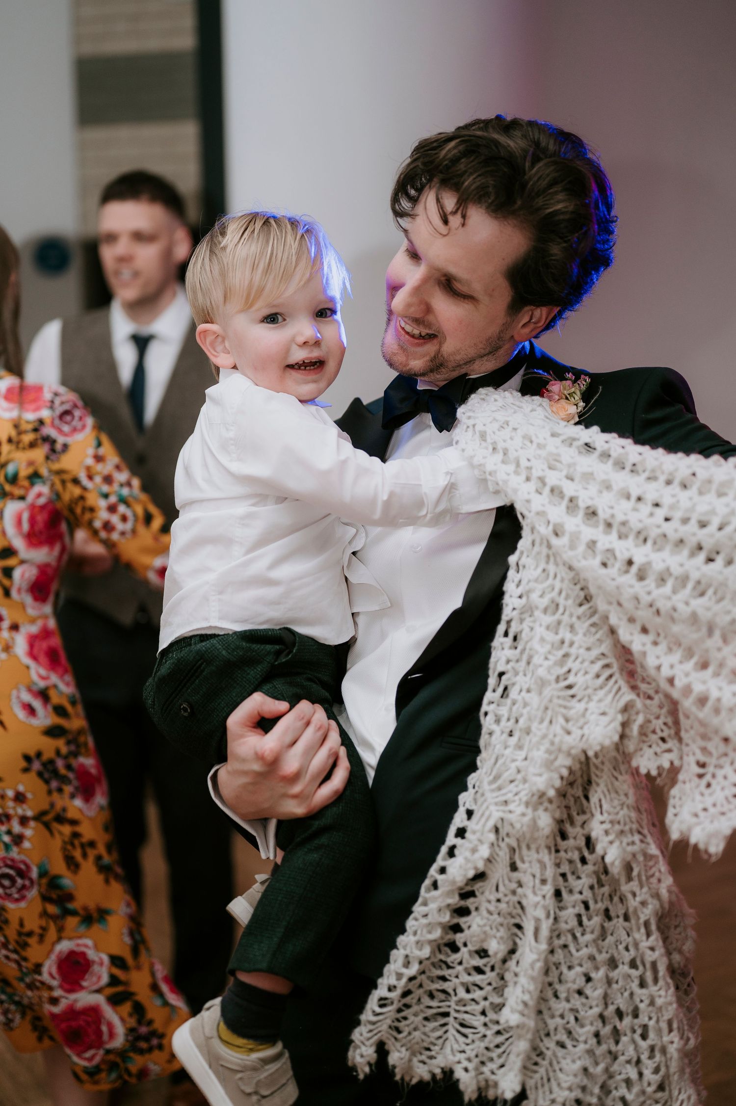 A toddler in white formal wear dances with a guest in a tuxedo at a wedding reception.