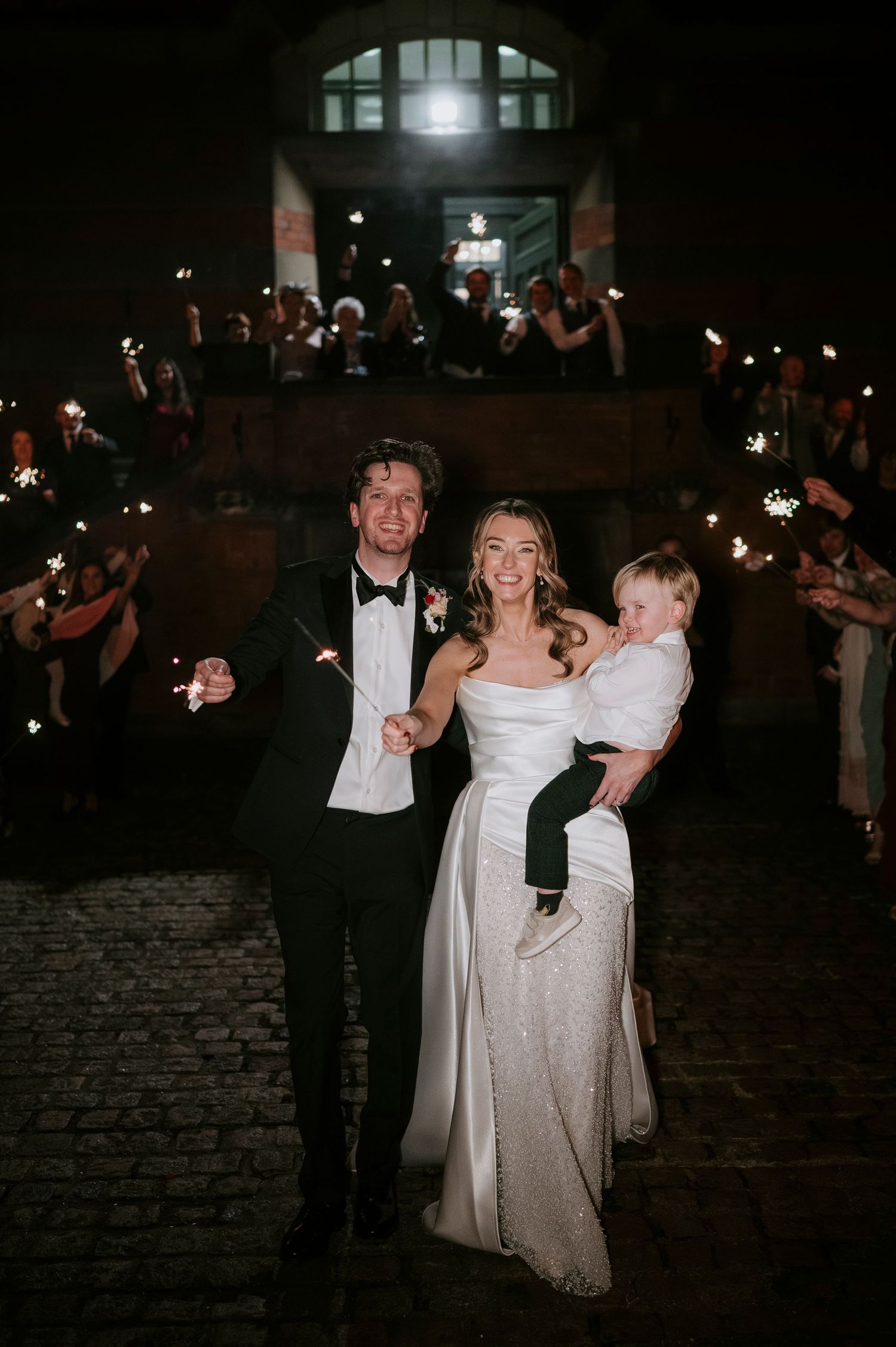 A couple dances joyfully at their wedding reception under twinkling lights on a cobblestone courtyard.
