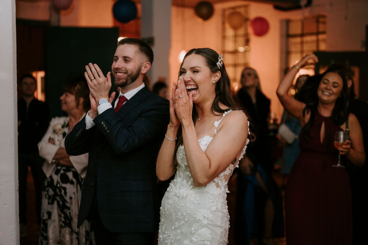 Excited bride in lace gown and groom in navy suit clap joyfully at their wedding reception.