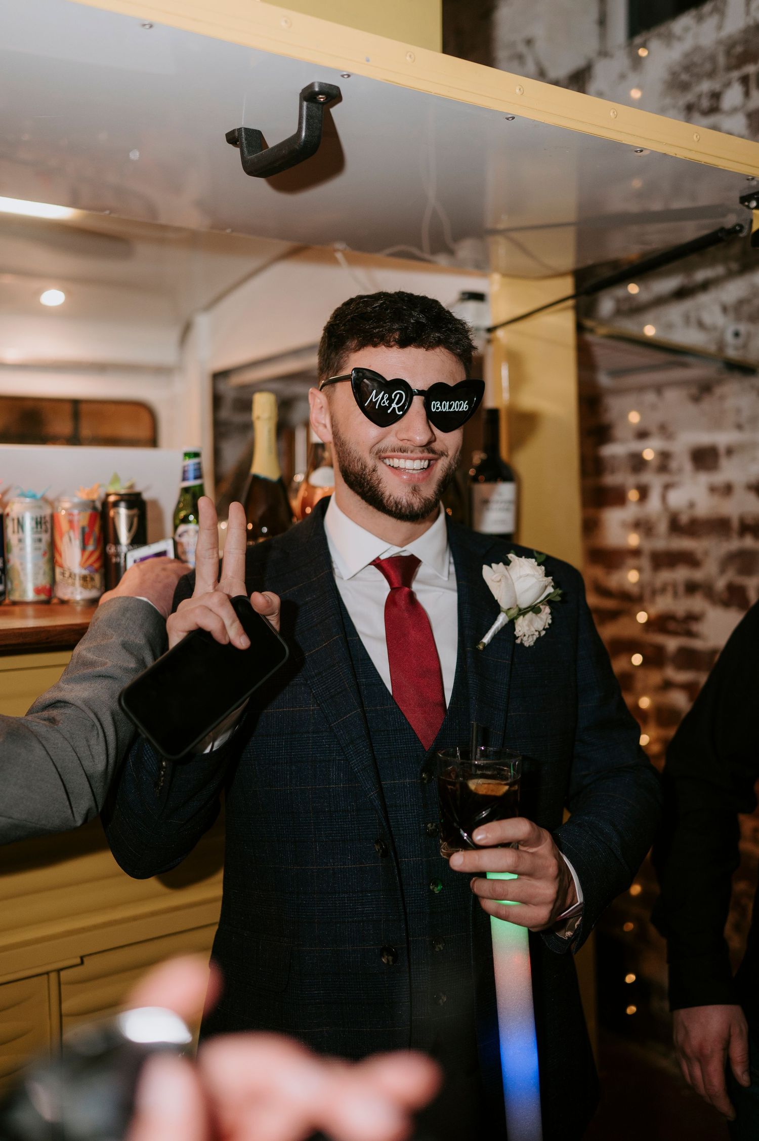 Smiling groom in navy suit wearing heart-shaped Mr & Mrs sunglasses holding drink at wedding bar.