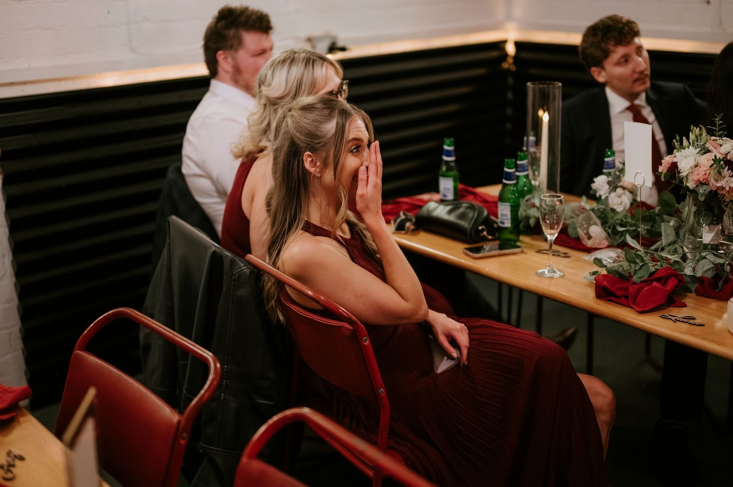 An emotional wedding guest in a burgundy dress covers her face while seated at a decorated reception dinner table.