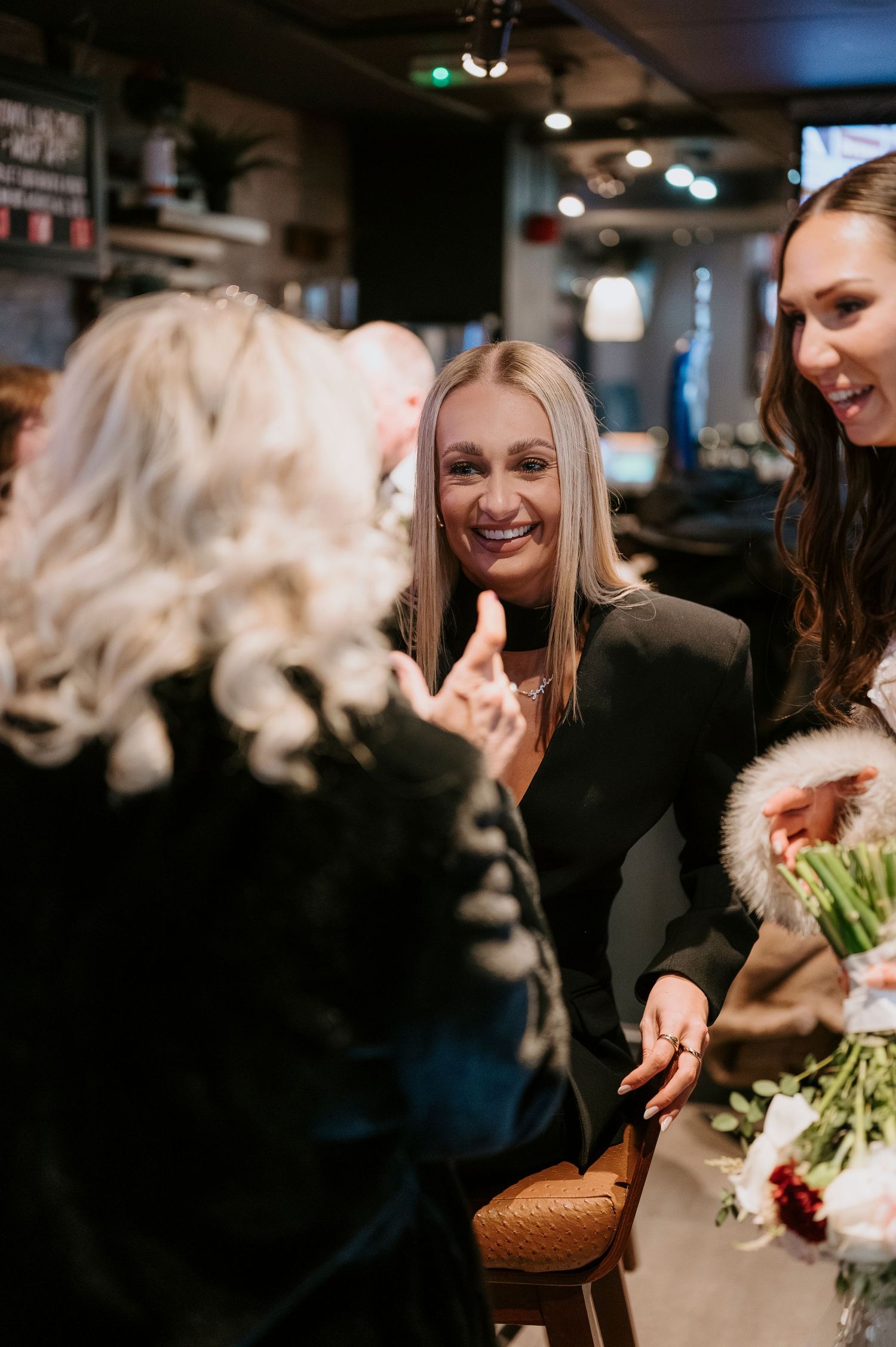 Blonde wedding guest in black blazer smiles warmly while chatting with bride and guests at reception.