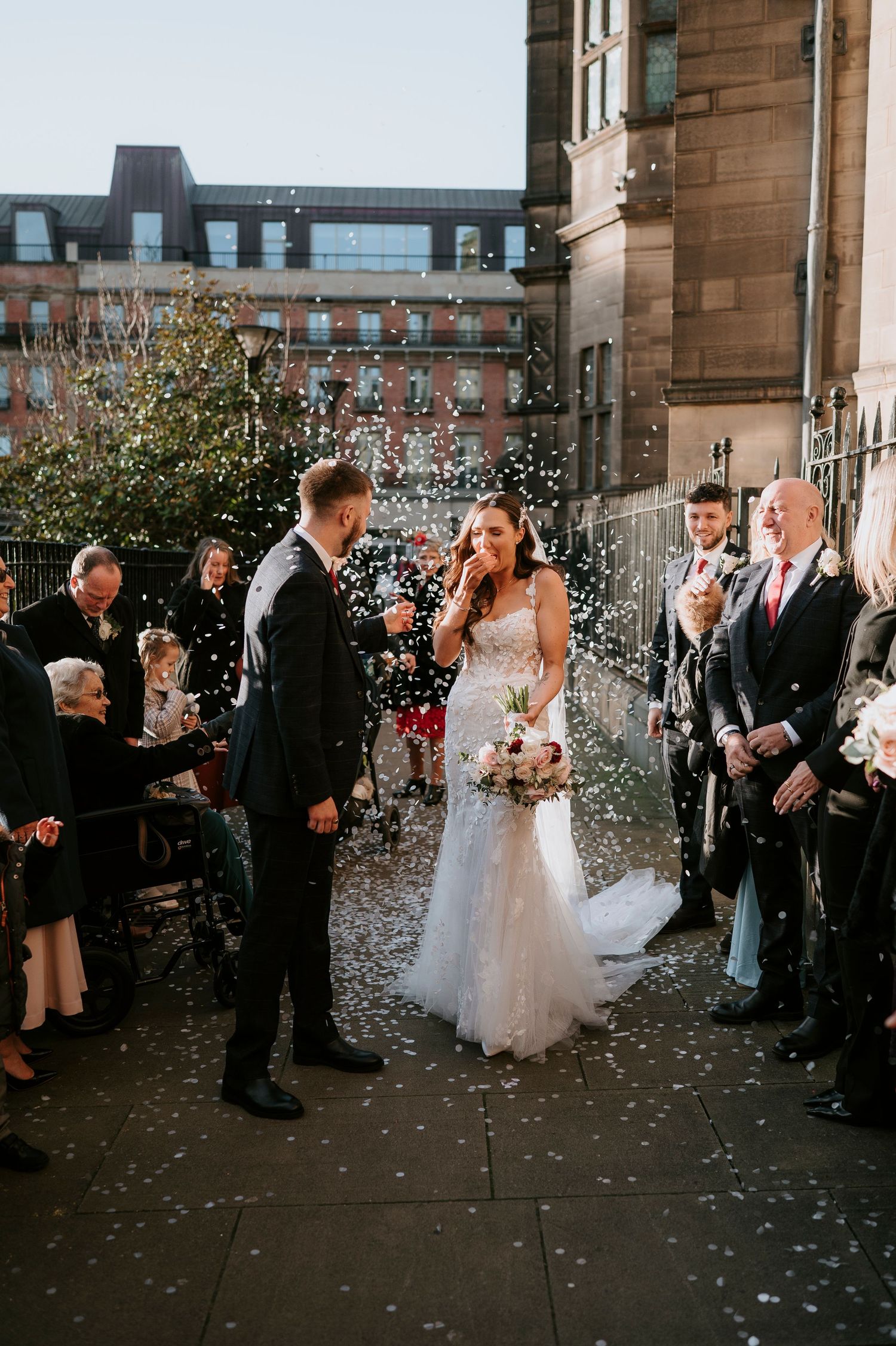 Bride and groom walk through confetti shower outside stone church surrounded by cheering wedding guests.
