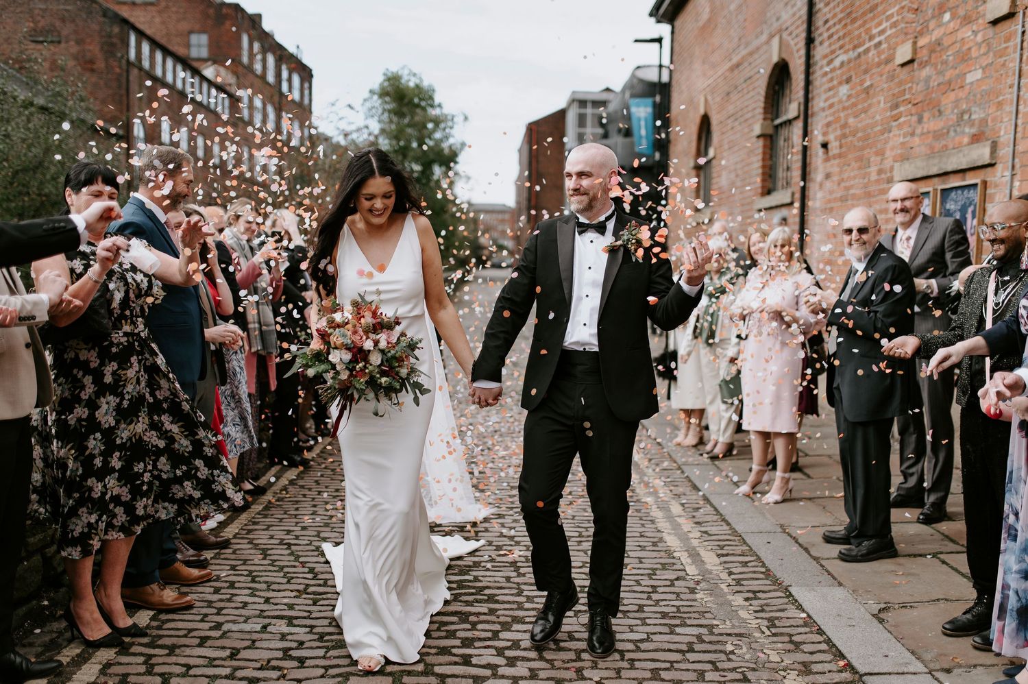 Newlyweds walk down cobblestone street while guests throw confetti during wedding celebration outside brick buildings.