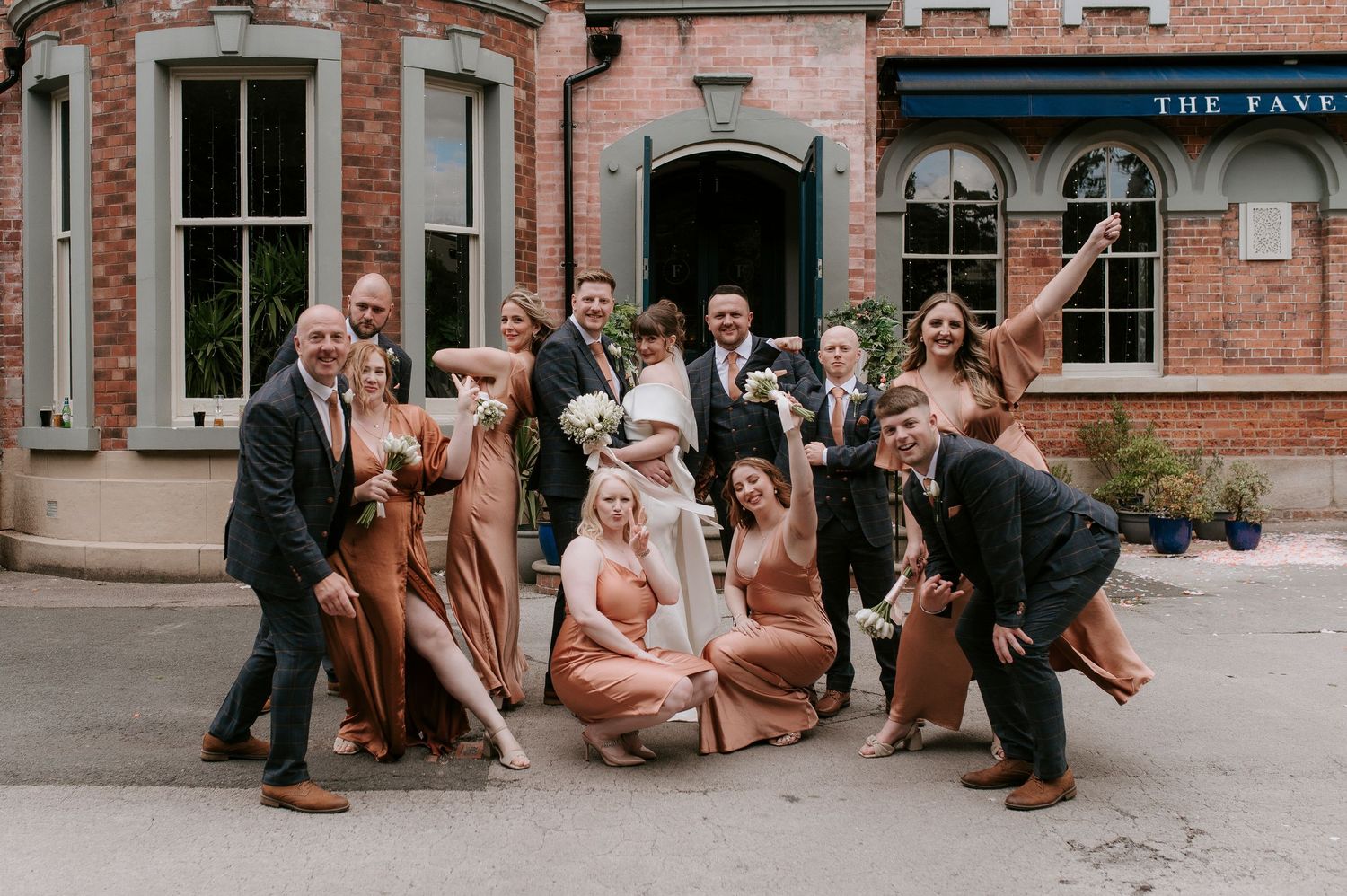 A wedding party poses playfully outside a brick building with vintage architecture.