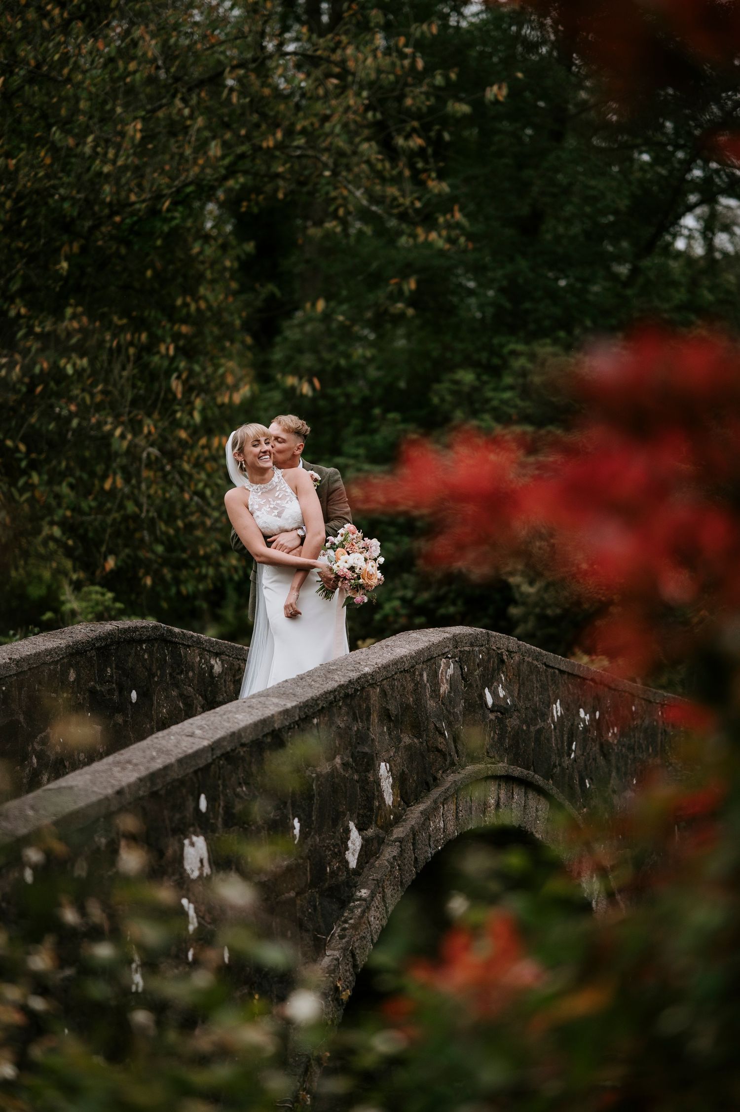 A couple stands embracing on a stone bridge surrounded by autumn foliage and red maple leaves in a romantic garden setting.
