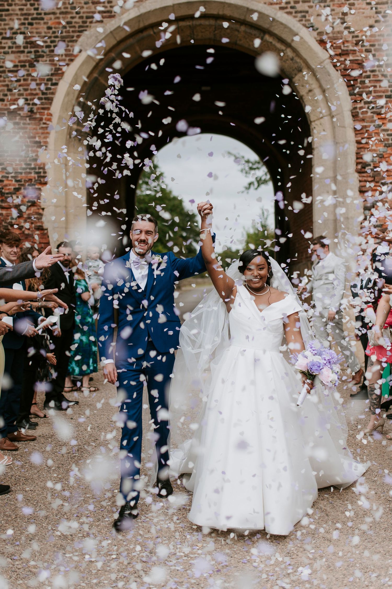 Wedding couple exits historic brick church archway while guests shower them with white confetti in joyful celebration.