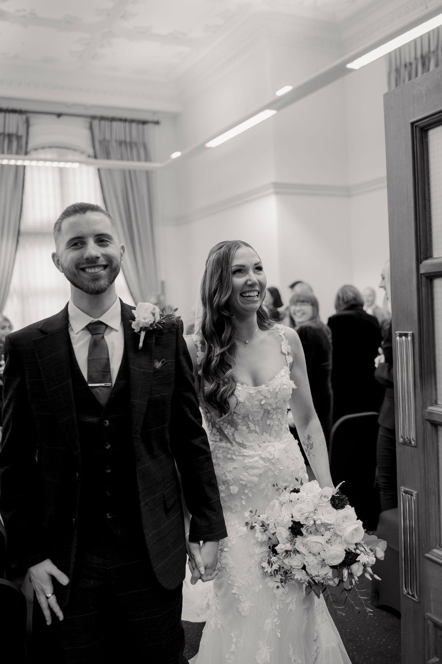 A wedding party walks and laughs together in a bright hallway.