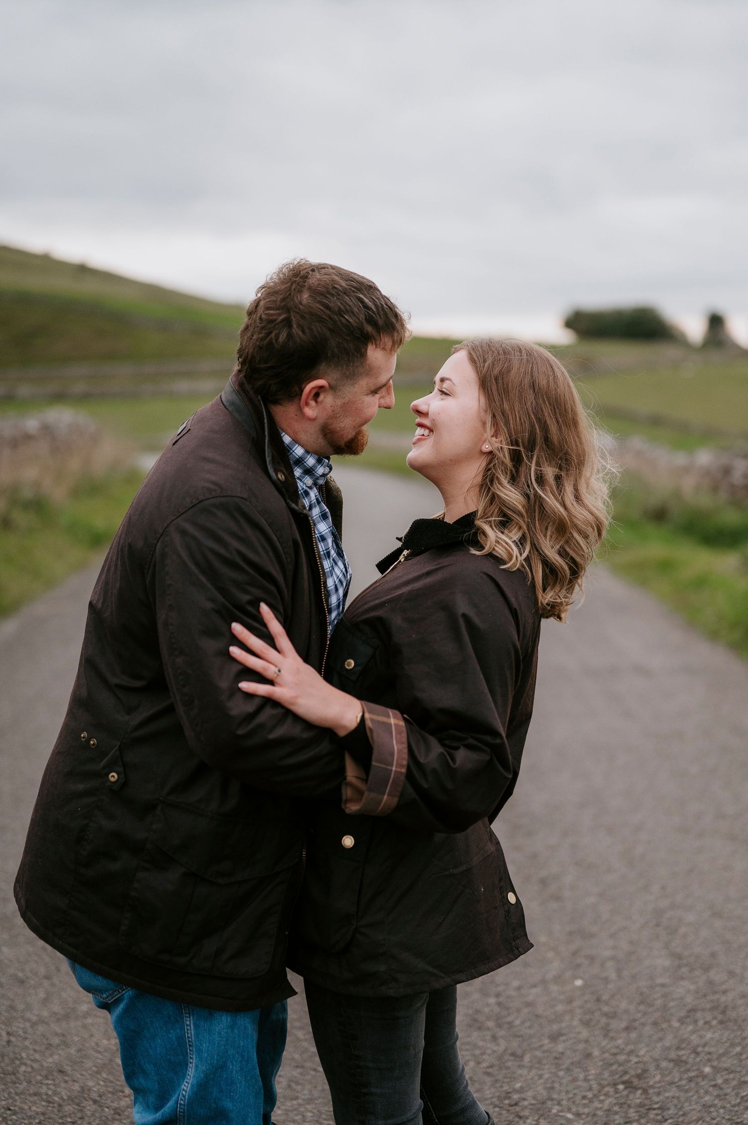 A couple in casual winter wear share intimate moments on a rural road under cloudy skies.