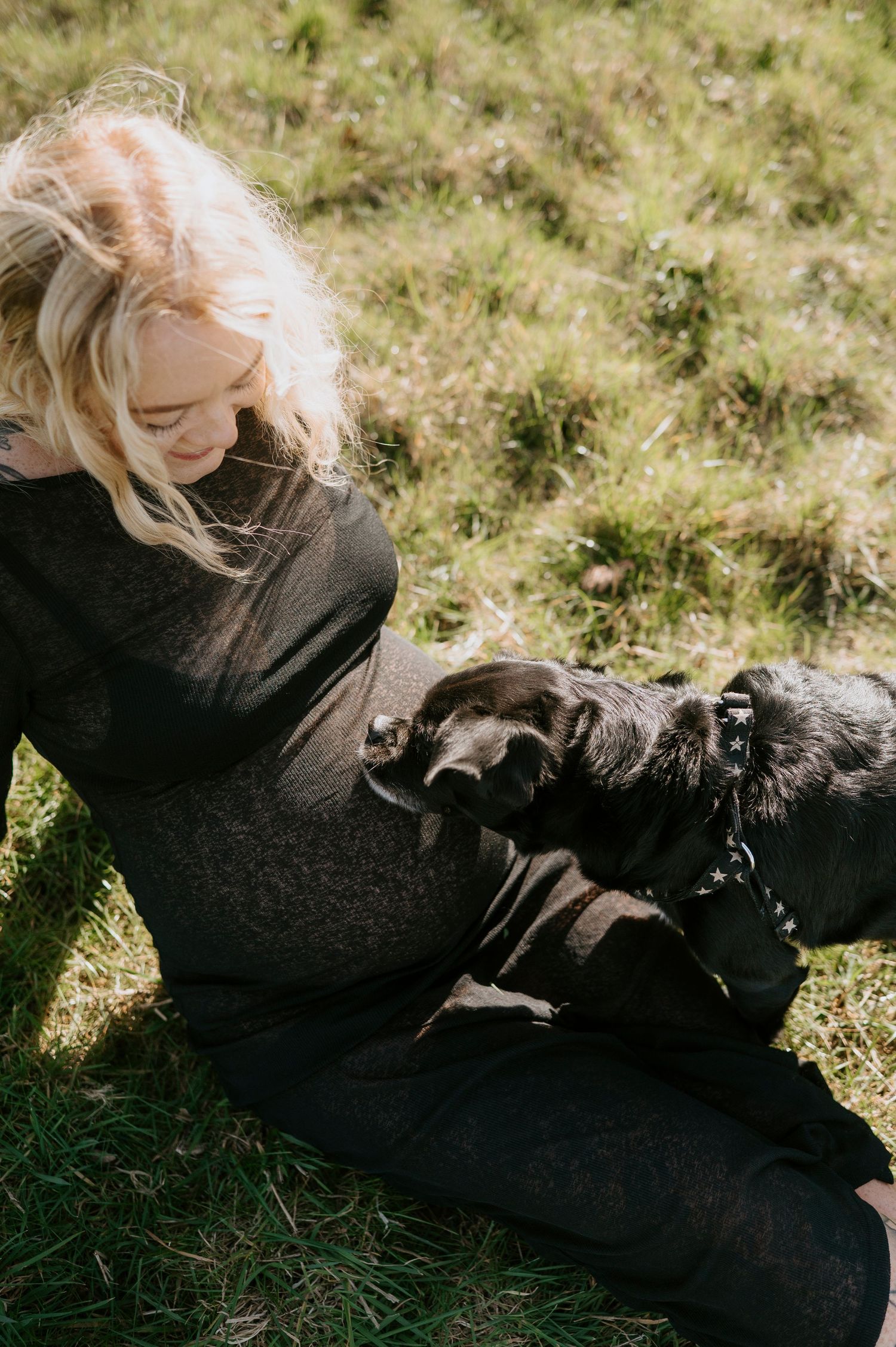 Maternity photos taken outdoors with a black dog joining the photoshoot on grassy ground.