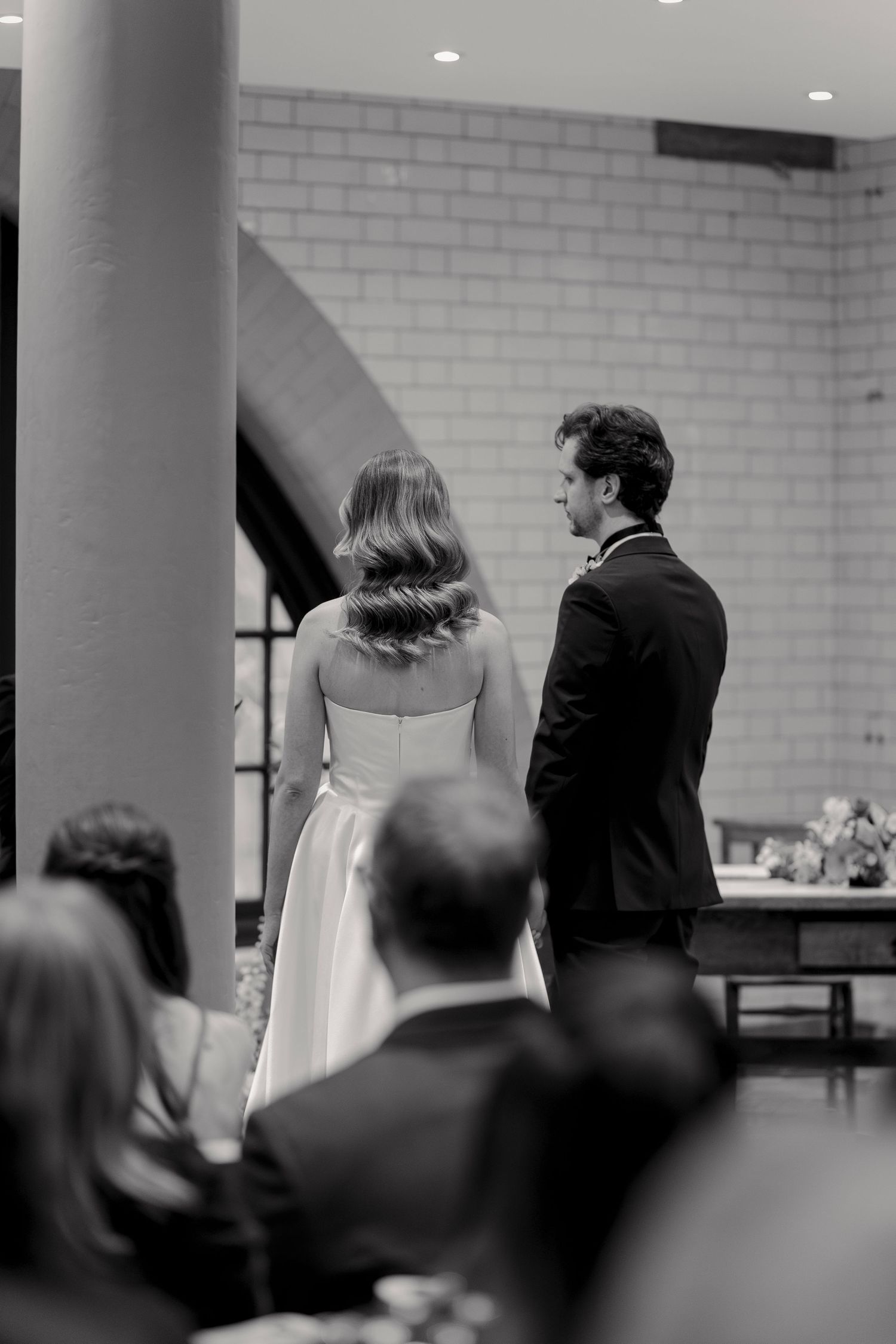 Black and white view of wedding ceremony guests seated in rows facing forward in an indoor venue.