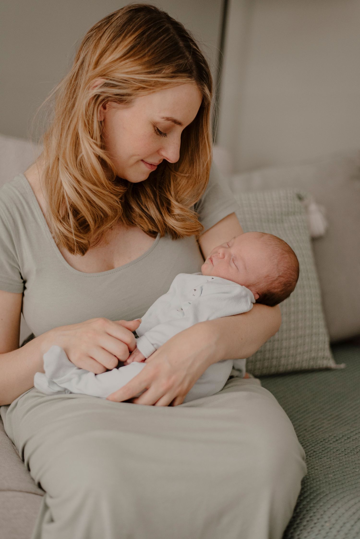 A tender moment as a mother in gray clothing cradles her sleeping newborn baby on a couch with throw pillows.