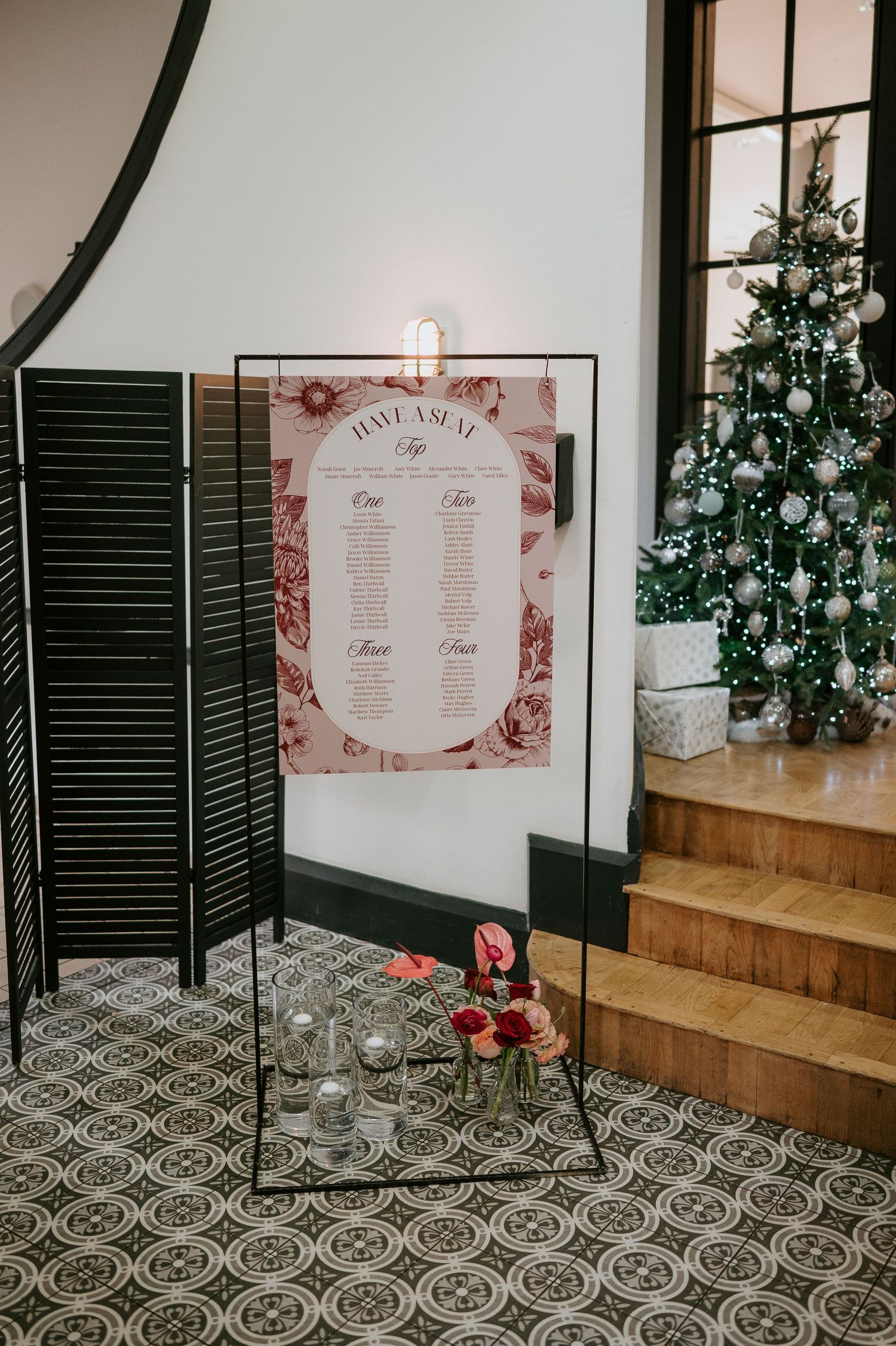 Wedding seating chart display with black shutters and floral decorations against a white wall.