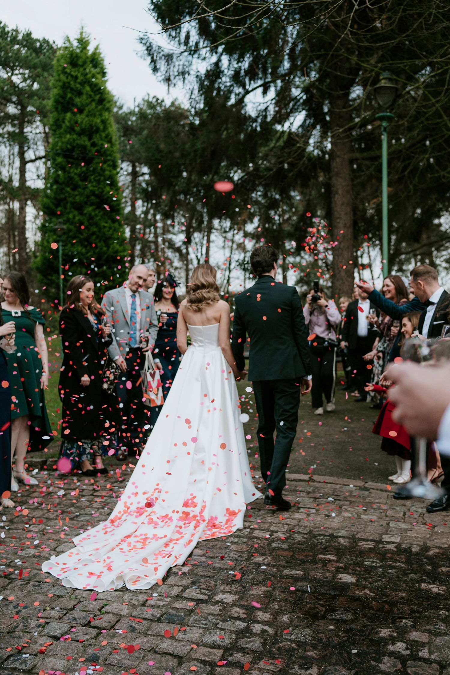 Wedding couple walks down path while guests shower them with pink and red rose petals under trees.