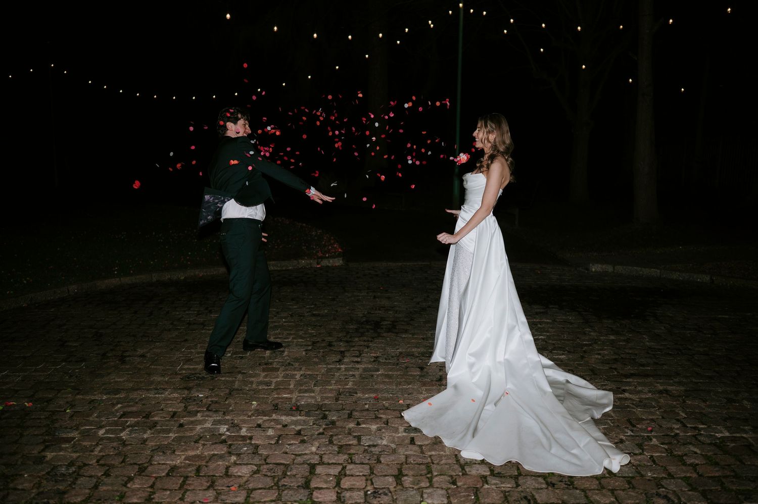 Couple dancing under twinkling string lights at night on a cobblestone patio during a wedding celebration.