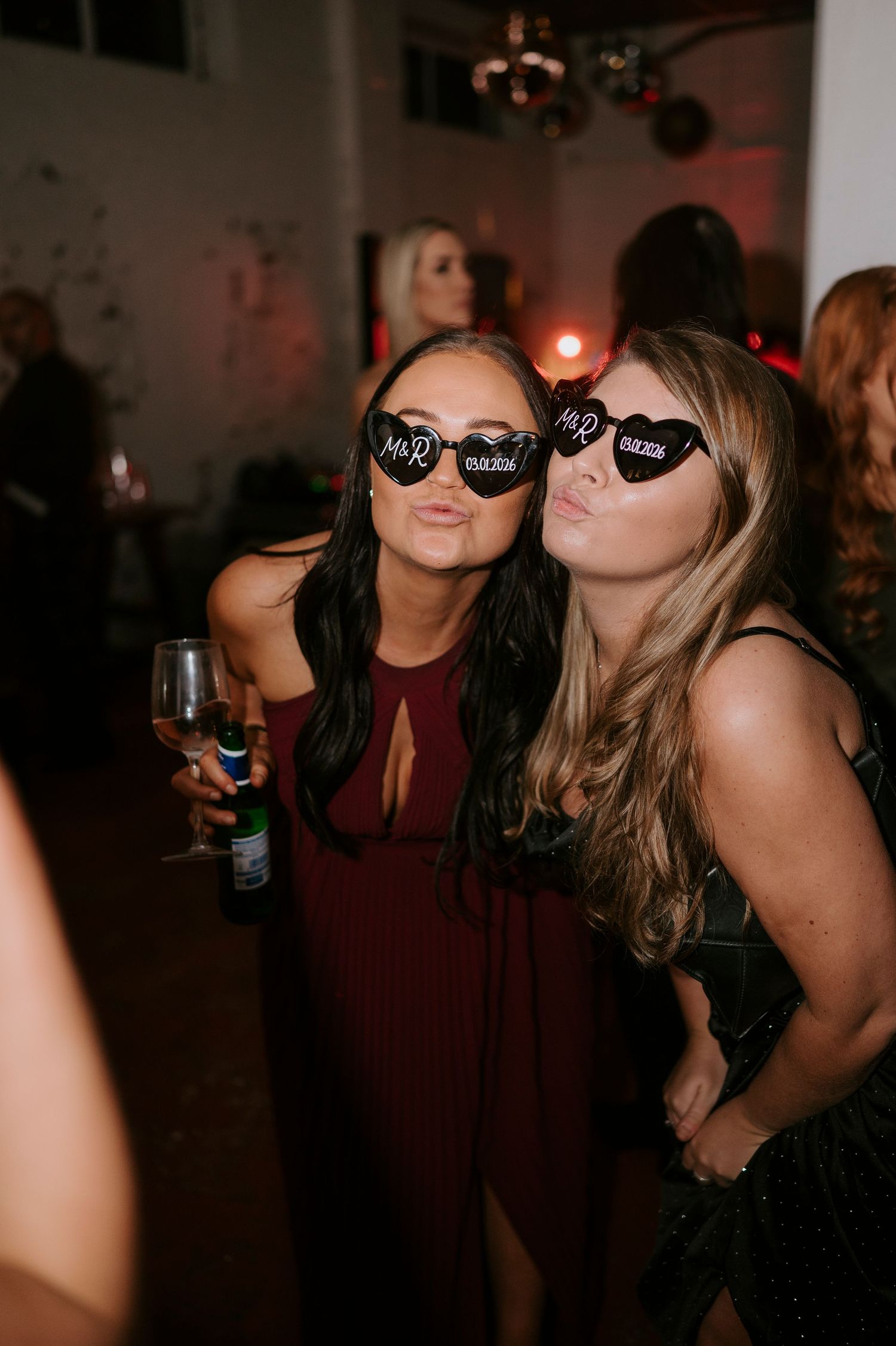Two women pose with heart-shaped Mr and Mrs sunglasses at a wedding reception party.