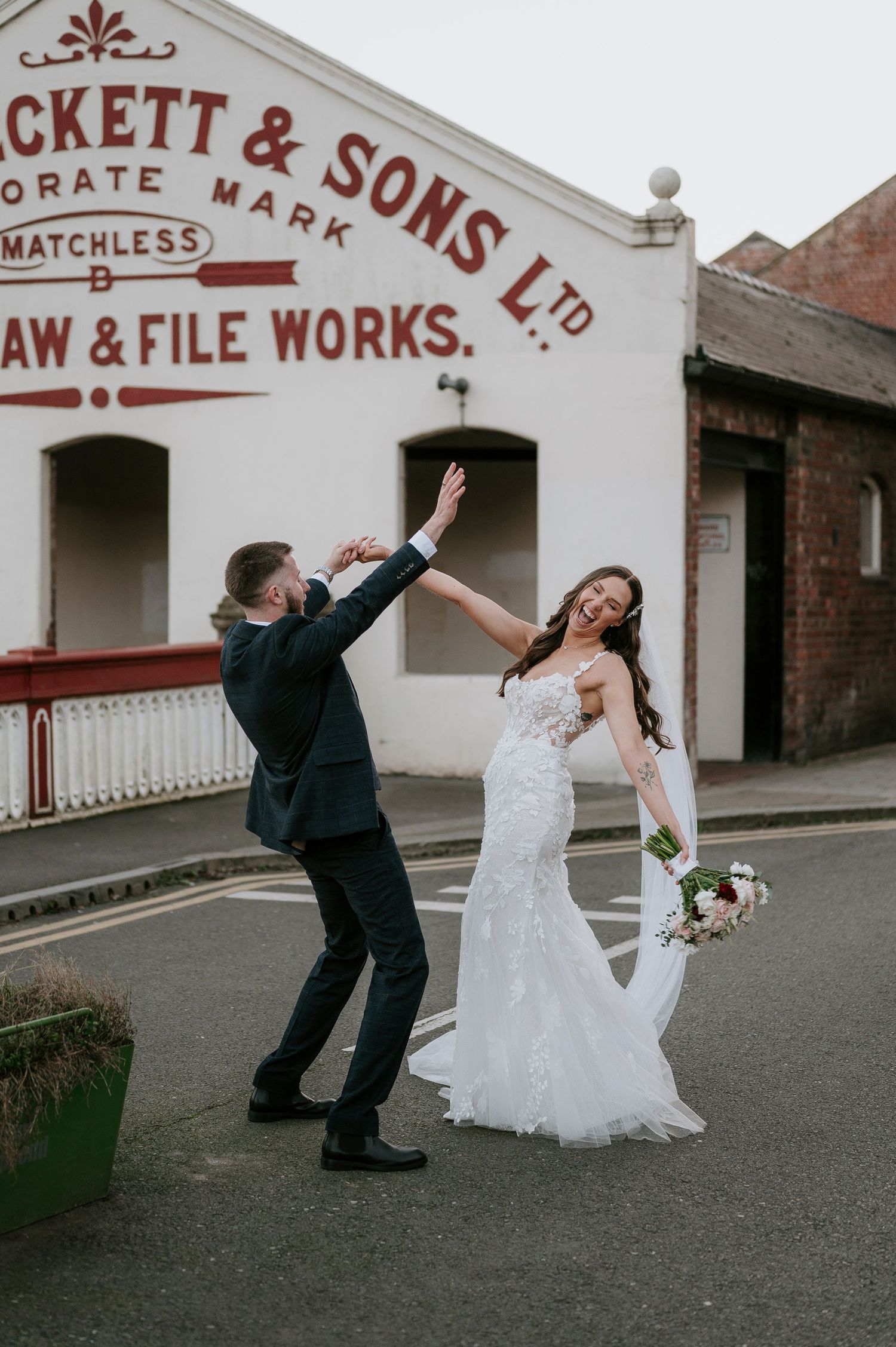 Groom twirls bride in lace wedding gown outside vintage Beckett & Sons Saw & File Works building.