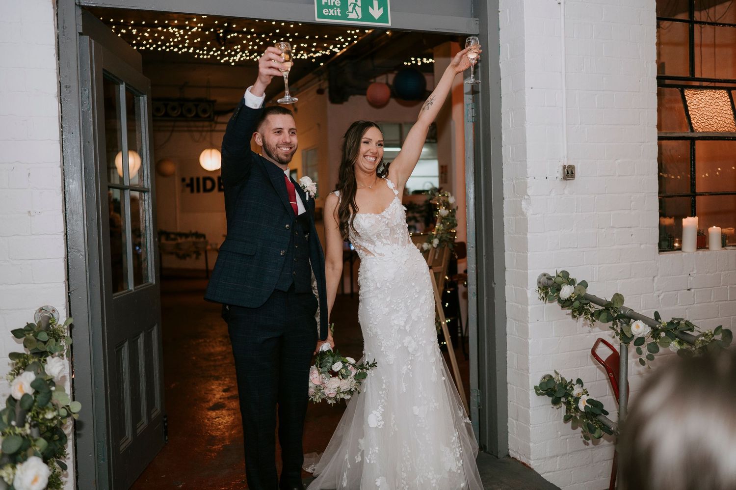 Smiling bride and groom raise hands in celebration as they enter their wedding reception through decorated doorway.