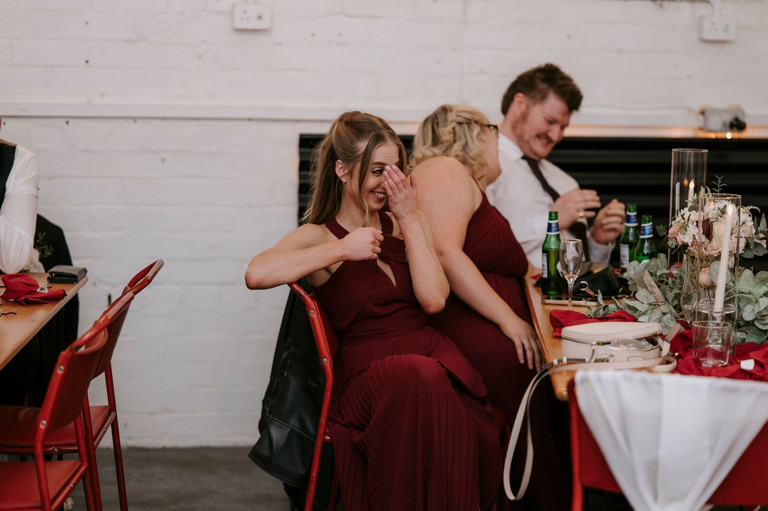 Two bridesmaids in burgundy dresses share a laugh at a wedding reception with red chairs and festive table decor.