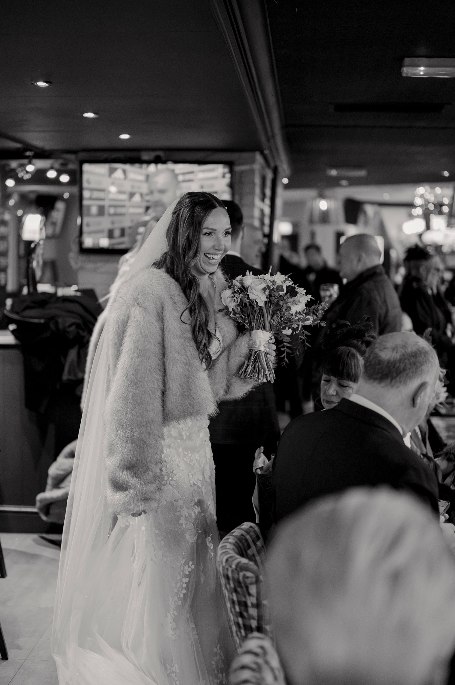 Smiling bride in lace gown and fur stole holds bouquet while greeting guests at wedding reception.